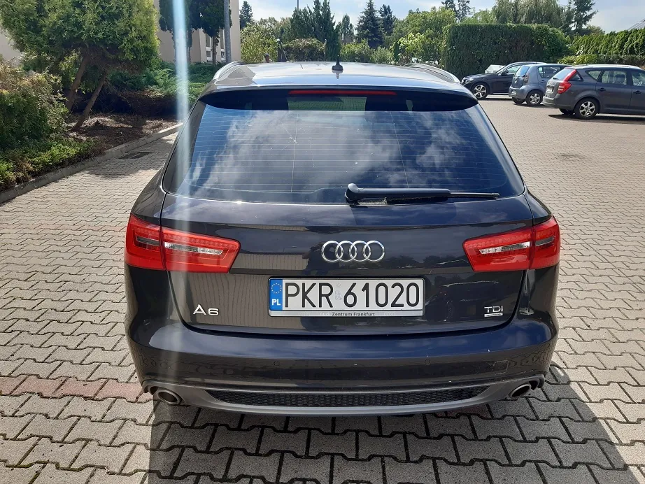 Tylny widok czarnego samochodu marki Audi A6 TDI zaparkowanego na kostce brukowej. Na samochodzie znajduje się polska tablica rejestracyjna o numerze PKR 61020. W tle widoczne są inne zaparkowane samochody, zieleń oraz budynki.