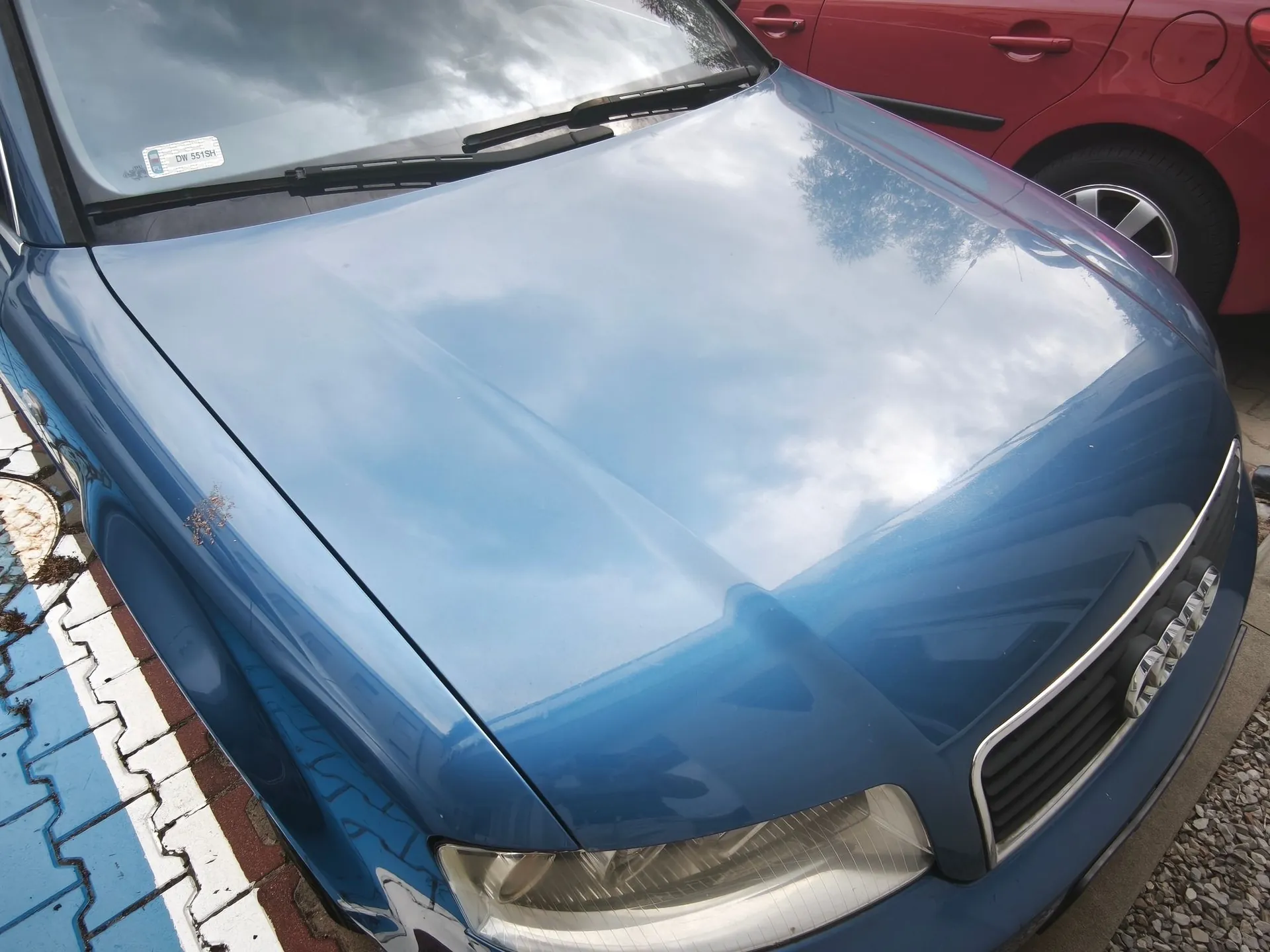 Zbliżenie na przednią maskę i reflektor niebieskiego samochodu marki Audi. Na masce widoczne są odbicia chmur i nieba oraz niewielkie uszkodzenie lakieru po lewej stronie. W tle widoczny jest fragment czerwonego samochodu oraz fragment chodnika z niebieskimi i białymi płytkami.