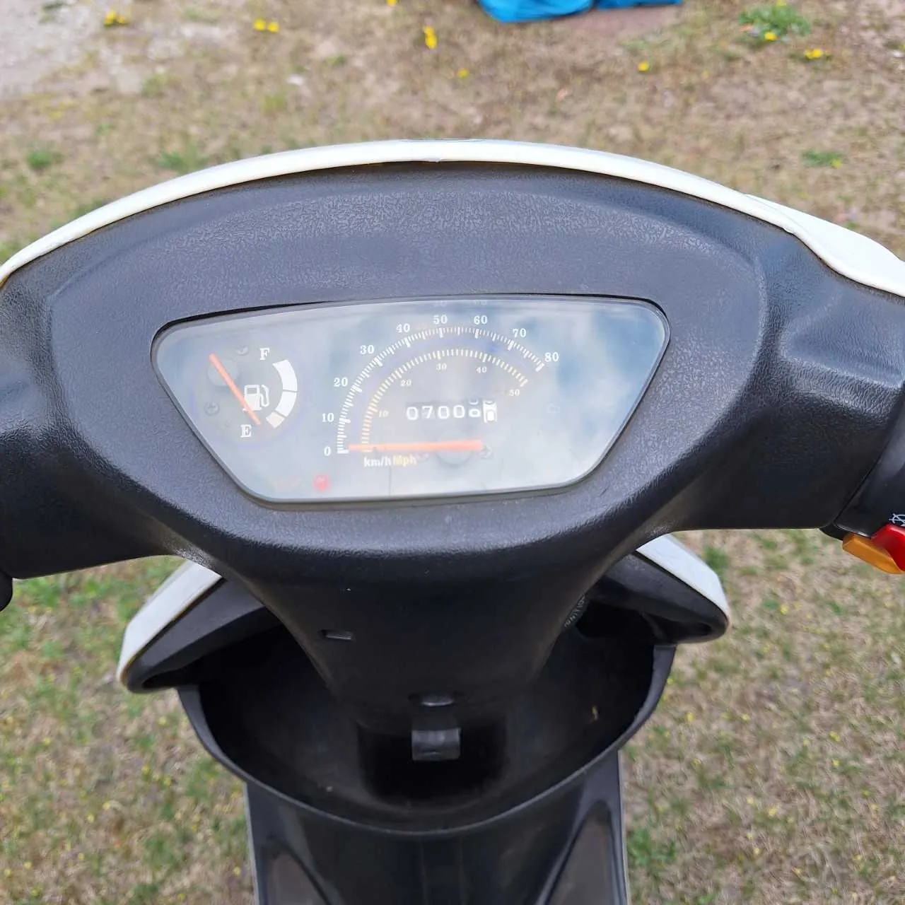 Zbliżenie na deski rozdzielcze skutera. Widoczny wskaźnik prędkości z zakresem 0-80 km/h, cyfrowy licznik przebiegu pokazujący 0700, wskaźnik paliwa z oznaczeniami E (pusty) i F (pełny), oraz napisy km/h Mph. Tło stanowi trawa z żółtymi kwiatami i niebieskim przedmiotem w oddali. Kierownica skutera jest czarna z białymi elementami, a po prawej stronie widoczny jest czerwony i pomarańczowy przycisk.