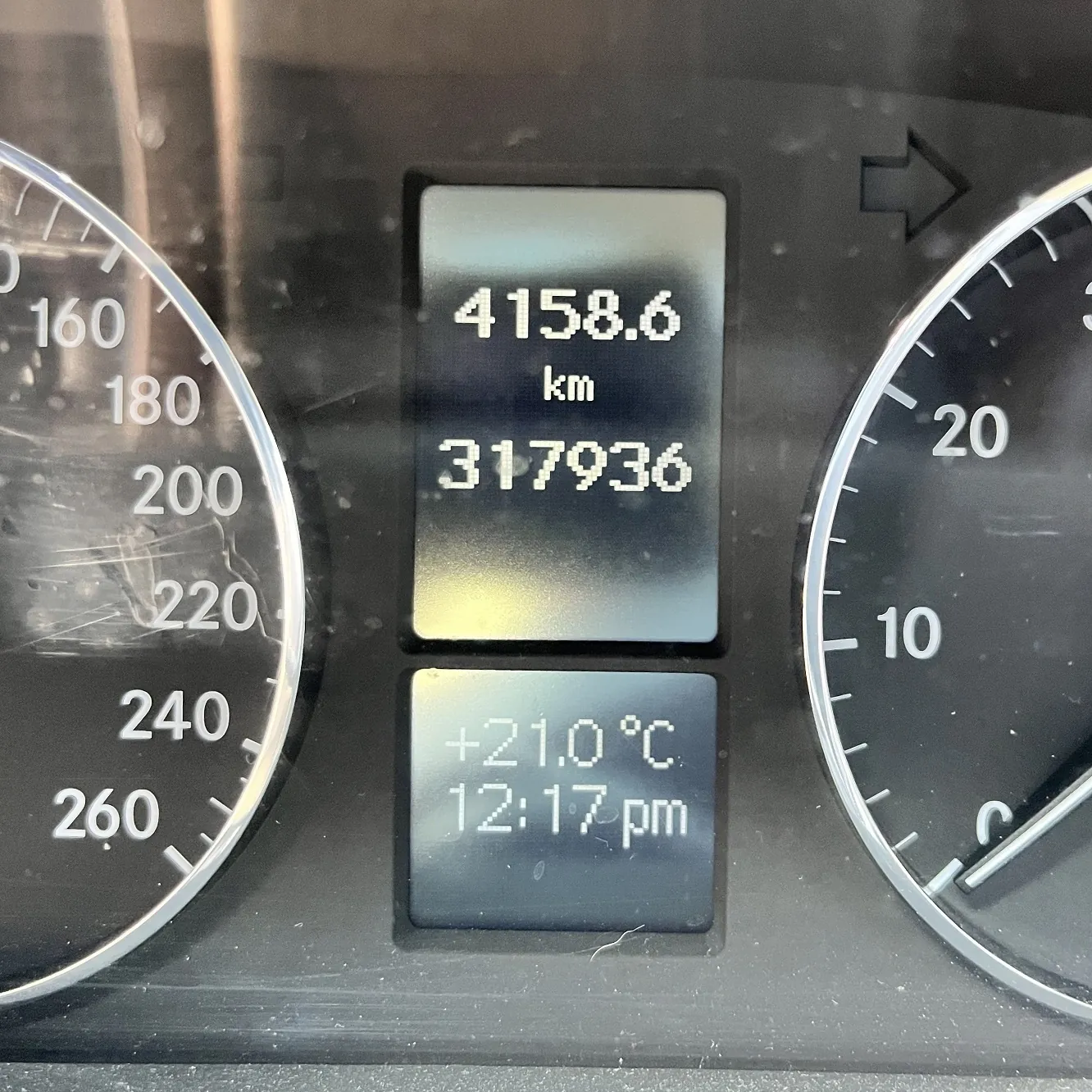 Zbliżenie na cyfrowy wyświetlacz deski rozdzielczej samochodu. Górny ekran pokazuje licznik dzienny 4158.6 km oraz całkowity przebieg 317936. Dolny ekran wyświetla temperaturę +21.0°C oraz godzinę 12:17 pm. Po bokach widoczne są fragmenty analogowych wskaźników prędkości i obrotów.