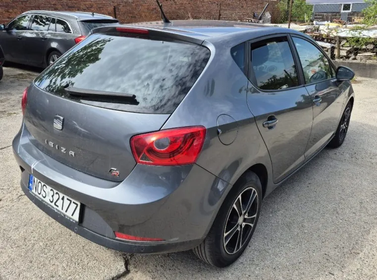 Szary samochód SEAT Ibiza z tyłu, widoczny napis IBIZA na bagażniku oraz tablica rejestracyjna NOS 32177. Po prawej stronie samochodu znajdują się czerwone światła tylnego. W tle drugi, ciemnoszary samochód, ceglana ściana oraz teren z żwirem. Samochód stoi na otwartym obszarze, w tle widoczne są budynki i roślinność.