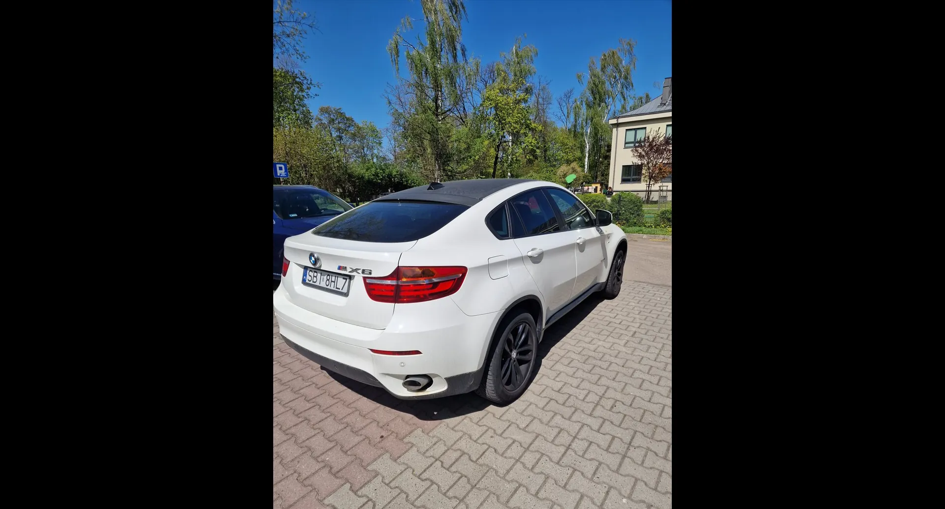 Biały samochód marki BMW X6 zaparkowany na kostce brukowej. Widok z tyłu i z boku pojazdu. Samochód ma czarny dach i czarne felgi. Tablica rejestracyjna to SBI 8HL7. W tle widoczne są zielone drzewa, fragment niebieskiego samochodu oraz jasny budynek mieszkalny pod bezchmurnym, niebieskim niebem.