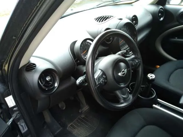 Wnętrze samochodu Mini Cooper z widocznym kierownicą z logo marki, czarnymi siedzeniami, szarą deską rozdzielczą z okrągłymi wentylatorami, wskaźnikami oraz dźwignią zmiany biegów. Drzwi po stronie kierowcy są otwarte, a na podłodze widoczne są pedały i dywanik. W tle przez przednią szybę widać fragment zewnętrzny.