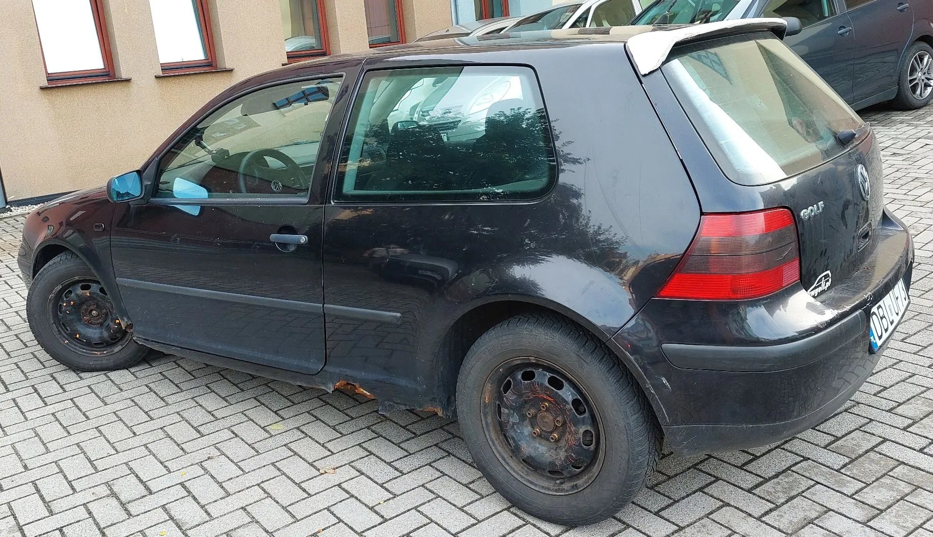 Czarny, trzy drzwiowy samochód Volkswagen Golf zaparkowany na kostce brukowej. Pojazd jest widoczny z boku i z tyłu, posiada stalowe felgi i widoczną rdzę na dolnej krawędzi progu między kołami. Z tyłu widoczny jest napis GOLF oraz fragment tablicy rejestracyjnej zaczynający się od liter DB.