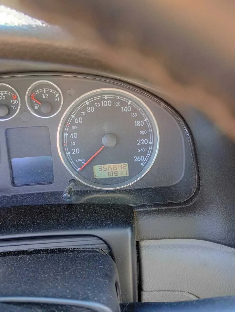 Zbliżenie na deski rozdzielcze samochodu. W centrum znajduje się prędkościomierz z zakresem do 260 km/h, wskazówka na 0 km/h. Na wyświetlaczu cyfrowym widoczny przebieg 356842 km oraz licznik dzienny 109.1 km. Po lewej stronie prędkościomierza znajdują się dwa mniejsze wskaźniki: termometr z zakresem 0-130°C oraz wskaźnik paliwa z oznaczeniami 1/2 i 1/1. Deska rozdzielcza jest zapylona, widoczna część kierownicy na górze oraz fragment panelu drzwiowego po prawej stronie.