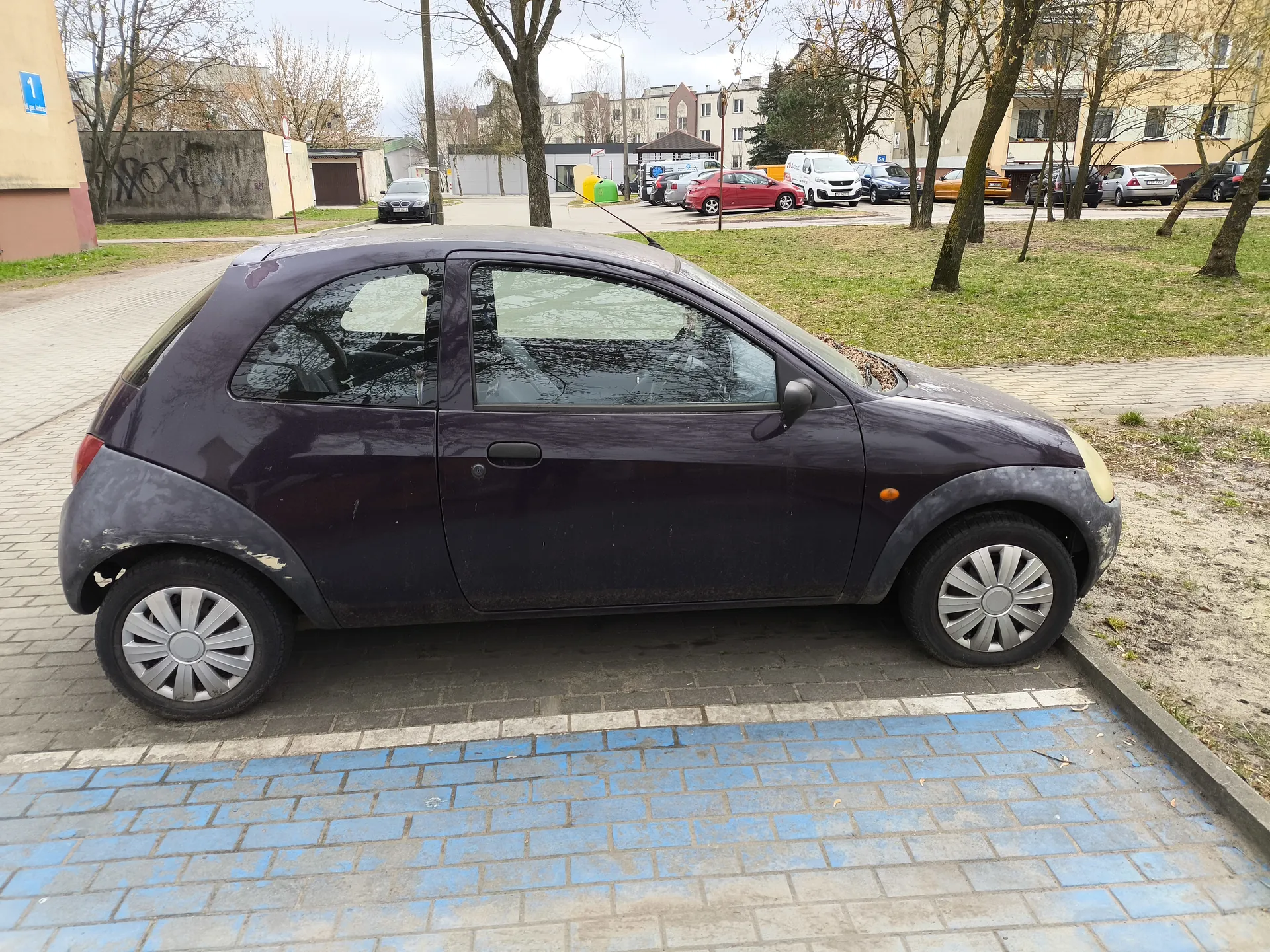 Ciemnofioletowy mały samochód osobowy zaparkowany na chodniku z niebieskimi i szarymi płytkami. Samochód ma srebrne felgi, zarysowane boki i brudną karoserię. W tle widoczne są inne zaparkowane pojazdy, drzewa bez liści, budynki mieszkalne, betonowa ściana z graffiti oraz niebieski znak z cyfrą 1 na budynku po lewej stronie. Niebo jest zachmurzone.