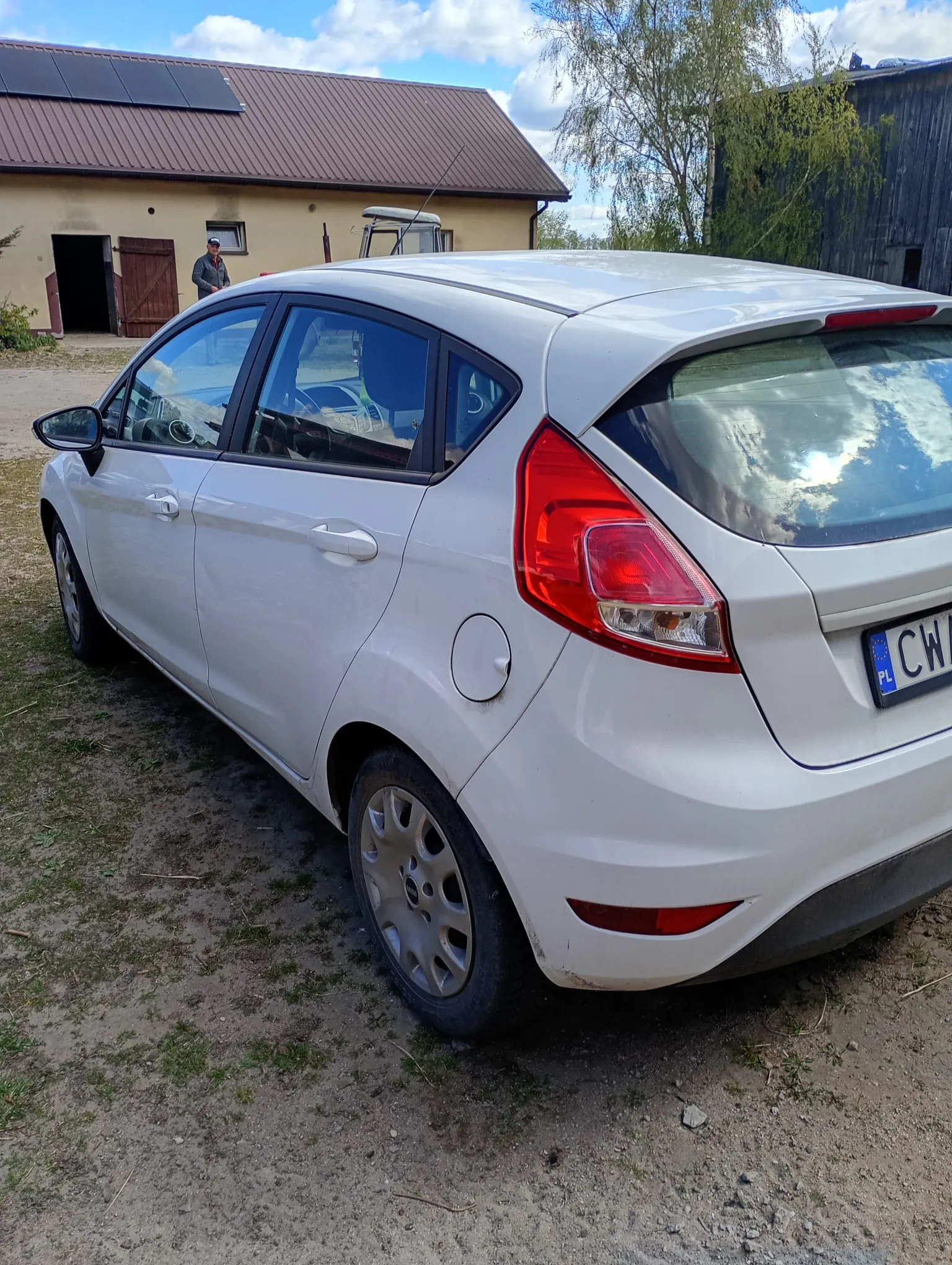 Biały samochód osobowy typu hatchback zaparkowany na podwórku z ziemią i trawą. Widoczna tylna część pojazdu z czerwonymi światłami i częściowo widoczną tablicą rejestracyjną z napisem PL CW. W tle budynek z brązowym dachem, panelami słonecznymi i otwartymi drzwiami, obok stoi osoba. Na niebie białe chmury.