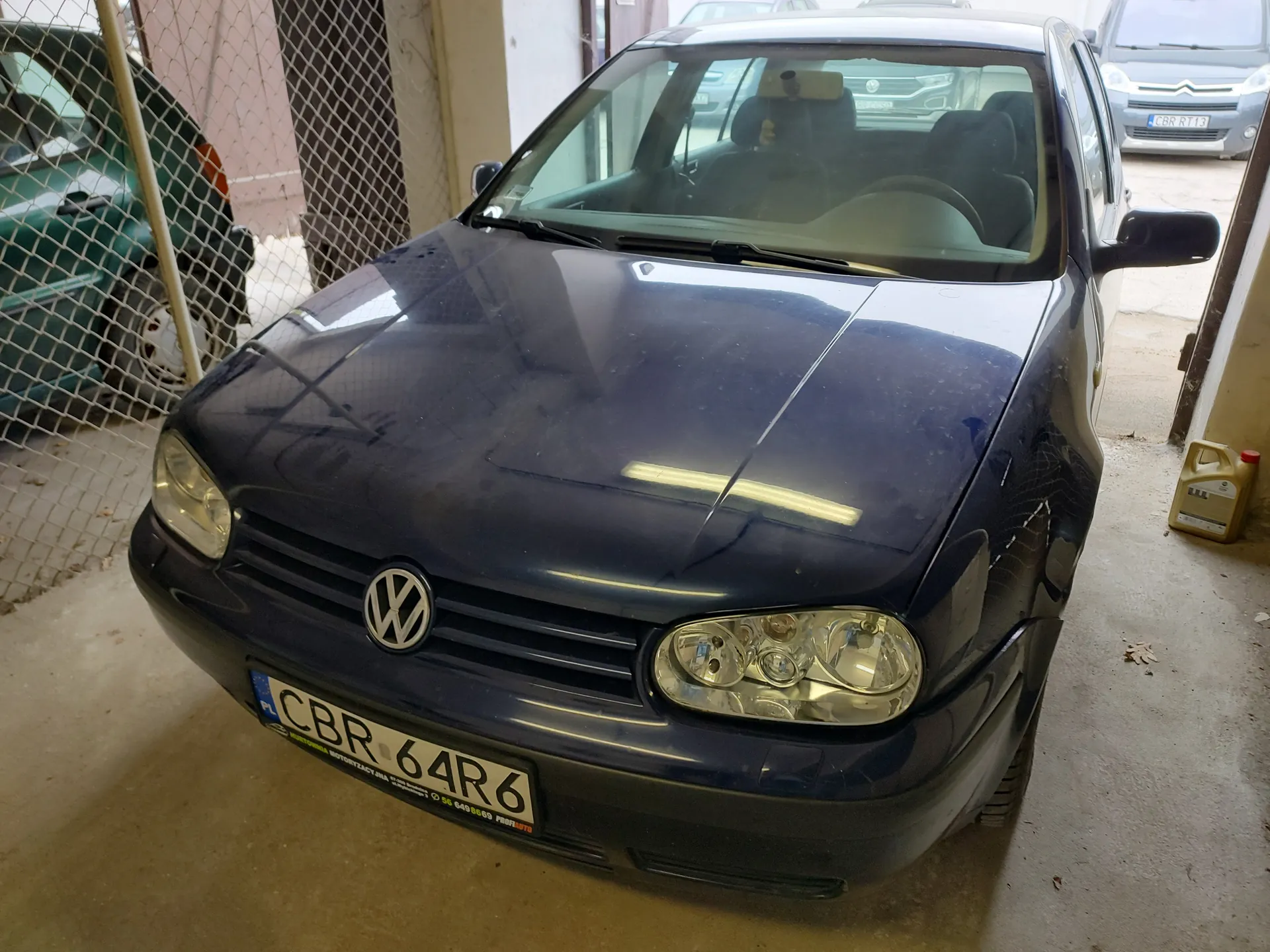 Ciemnoniebieski samochód marki Volkswagen z tablicą rejestracyjną CBR 64R6 (PL) zaparkowany w garażu. W tle widoczne są inne pojazdy, w tym zielony samochód za siatką oraz srebrny Citroën z tablicą CBR RT13. Na podłodze znajduje się pojemnik na olej. Wnętrze garażu z betonową podłogą i metalową siatką oddzielającą przestrzeń.