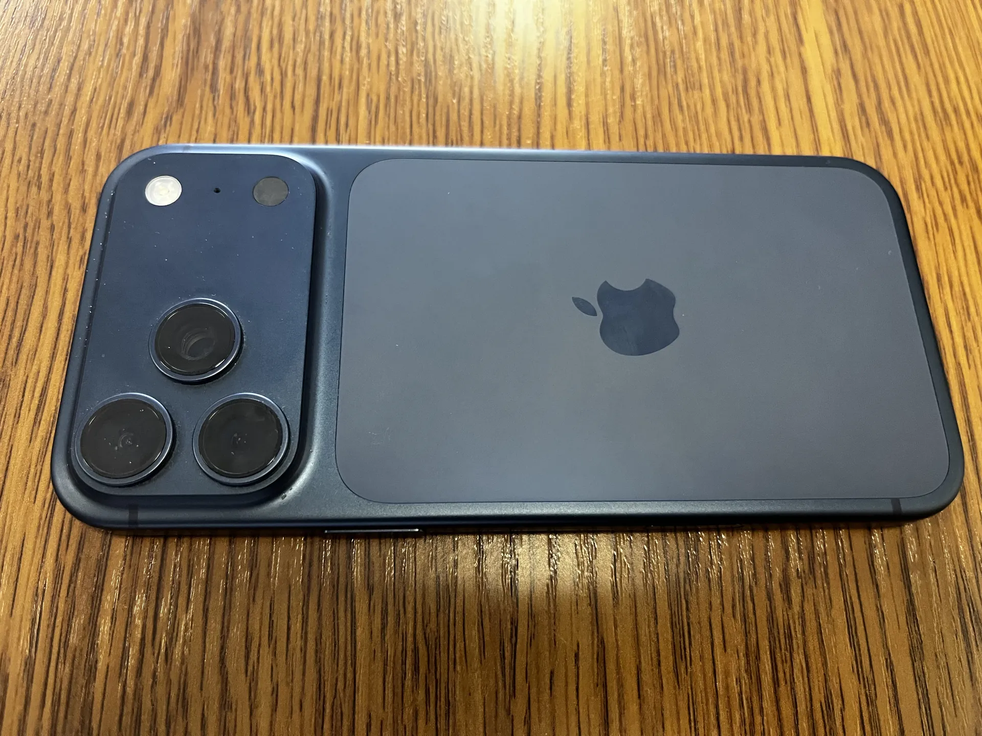 Zdjęcie przedstawia tylną stronę ciemnoszarego smartfona z logo Apple umieszczonym w centralnej części. Po lewej stronie znajduje się moduł z trzema okrągłymi obiektywami aparatu oraz dwoma mniejszymi okrągłymi elementami. Telefon leży na drewnianej powierzchni o widocznych pionowych żyłkach. Brak czytelnego tekstu na obrazie.