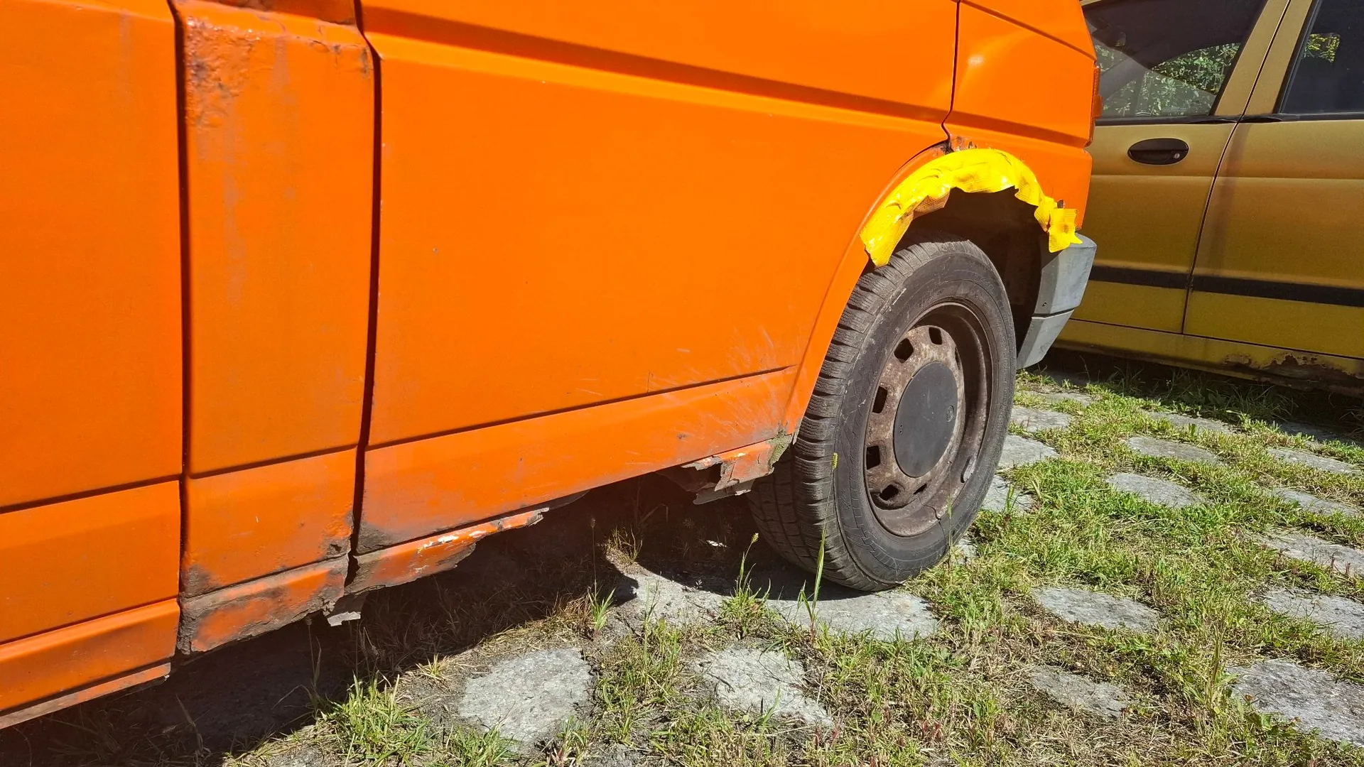 Zbliżenie na dolną część pomarańczowego samochodu. Widoczna jest tylna opona oraz dolna krawędź nadwozia z wyraźnymi śladami korozji i ubytkami w karoserii. Nad nadkolem znajduje się żółta taśma klejąca. Samochód stoi na podłożu z betonowych płyt z przerostami trawy. W tle widoczny jest fragment innego, żółtego pojazdu.