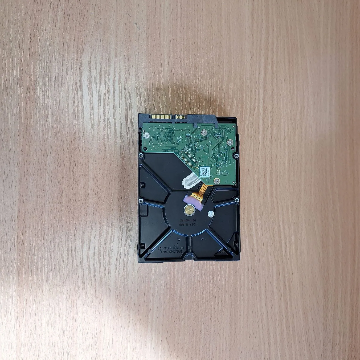 Zdjęcie przedstawiające spód dysku twardego HDD leżącego na jasnym, drewnianym blacie. Widoczna jest zielona płytka drukowana z komponentami elektronicznymi, metalowa obudowa oraz złącza SATA. Na obudowie znajduje się nadruk z numerami seryjnymi i oznaczeniami: '1257-A1880', '53 745 1794' oraz 'SATA-III 6Gb/s'.