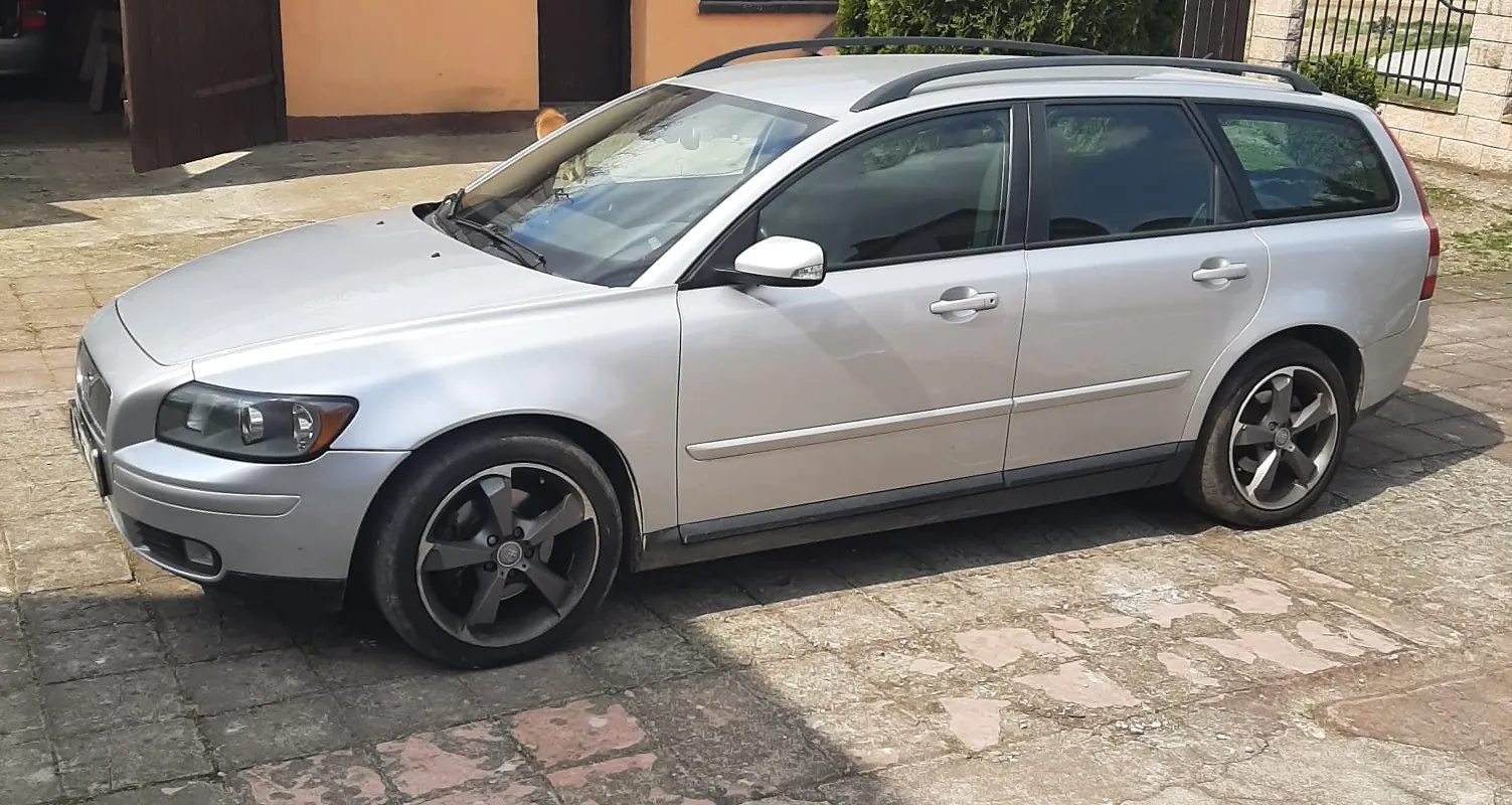 Srebrny samochód typu kombi z czarnymi felgami i dachowymi prętami, zaparkowany na brukowanej powierzchni. W tle pomarańczowa ściana budynku z brązowymi drzwiami, zielone krzewy oraz metalowe ogrodzenie. Samochód ma czarne opony, boczne lusterka i rękojeści drzwi w kolorze nadwozia.