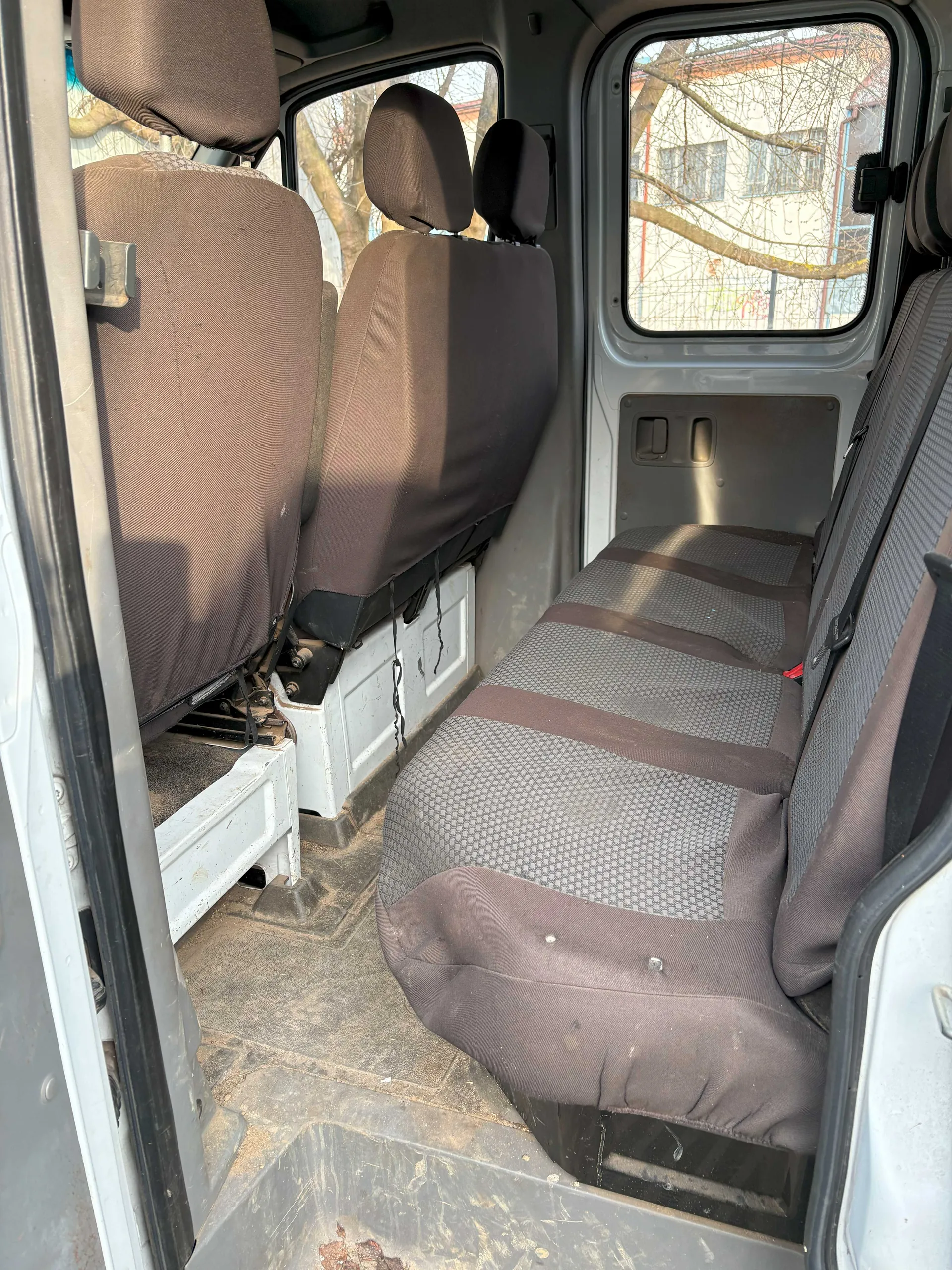 Wnętrze furgonetki z brązowymi pokrowcami na siedzeniach. Widoczne są trzy rzędy siedzeń, z których część jest przesunięta lub demontowana. Podłoga jest zabrudzona, a przez okna widać drzewa i budynek z siatką. Drzwi furgonetki są otwarte, a wnętrze oświetlone jest światłem dziennym.