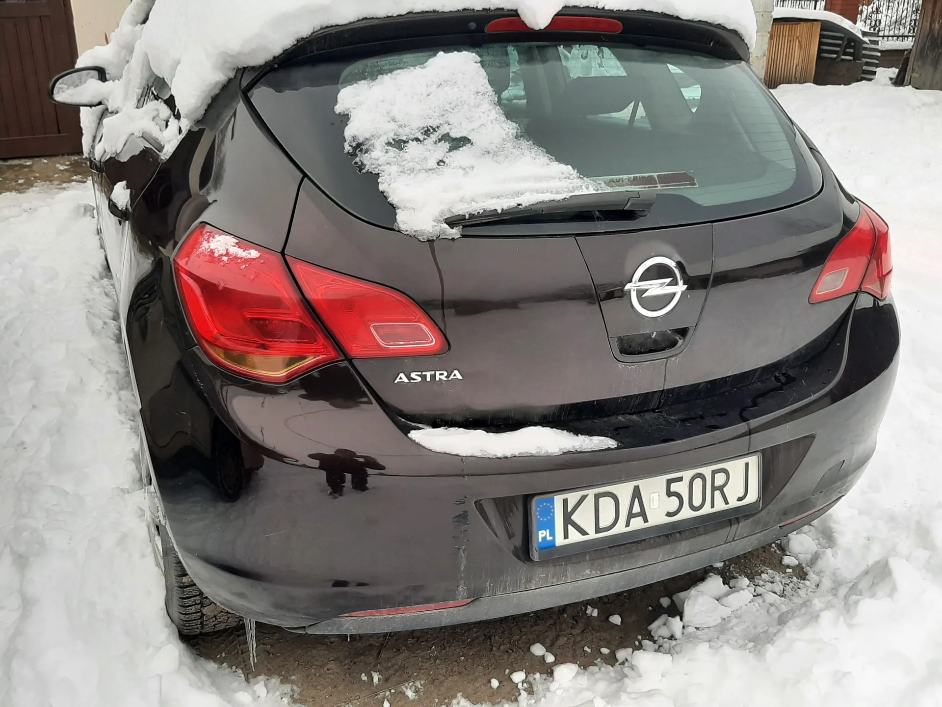 Czarny samochód Opel Astra z widocznym logo i napisem "ASTRA" na bagażniku. Na tylnej szybie i dachu leży śnieg. Tablica rejestracyjna KDA 50RJ z kodem "PL". Śnieżna sceneria, widoczne ślady opon i otoczenie z budynkami.