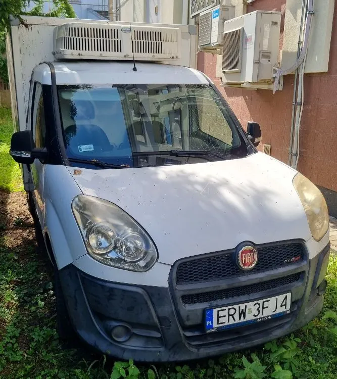 Biały samochód dostawczy marki Fiat z zabudową chłodniczą na dachu. Pojazd zaparkowany jest na trawie obok budynku, na którym zamontowane są dwie jednostki zewnętrzne klimatyzacji. Samochód posiada polską tablicę rejestracyjną o numerze ERW 3FJ4.
