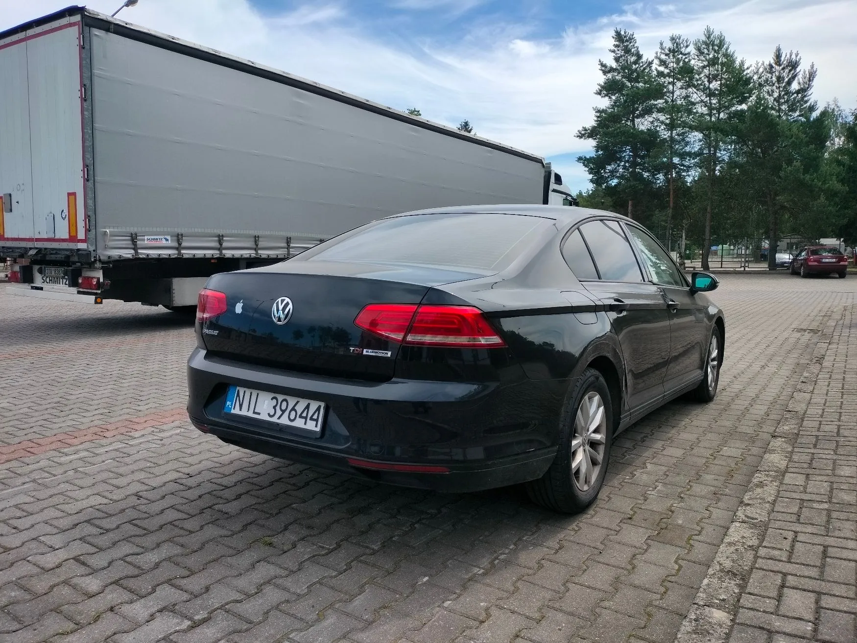 Czarny samochód osobowy marki Volkswagen zaparkowany tyłem do widza na kostce brukowej. Na klapie bagażnika widoczne jest logo Volkswagena oraz tablica rejestracyjna o numerze NIL 39644. W tle, po lewej stronie, znajduje się duża, biała naczepa ciężarówki marki Schmitz. Po prawej stronie widoczne są zielone drzewa i fragment innego czerwonego samochodu w oddali.