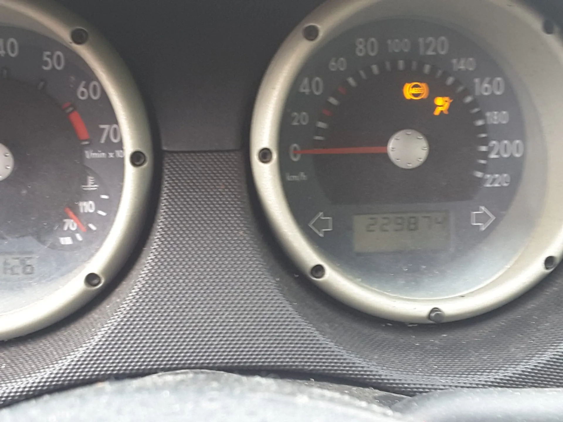 Zbliżenie na deskę rozdzielczą samochodu z dwoma okrągłymi zegarami. Po prawej stronie znajduje się prędkościomierz z podziałką od 0 do 220 km/h, wskazówka wskazuje 0. Na tarczy prędkościomierza świecą się dwie pomarańczowe kontrolki ostrzegawcze. Poniżej znajduje się cyfrowy licznik przebiegu wyświetlający wartość 229874. Po lewej stronie widoczny jest fragment obrotomierza z podziałką do 110 x 10 1/min.