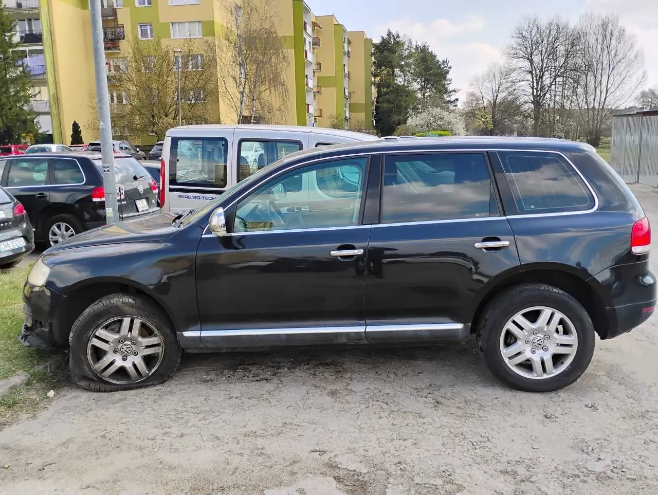 Czarny SUV marki Volkswagen z płaskim przednim lewym kołem zaparkowany na żwirowym placu. W tle widoczne są inne pojazdy, w tym biały furgon z napisem "FREMO-TECH", czarny samochód osobowy oraz bloki mieszkalne w kolorze żółto-zielonym. Na pierwszym planie sucha trawa i drzewa bez liści. Horyzont jest poziomy, budynki stoją pionowo.