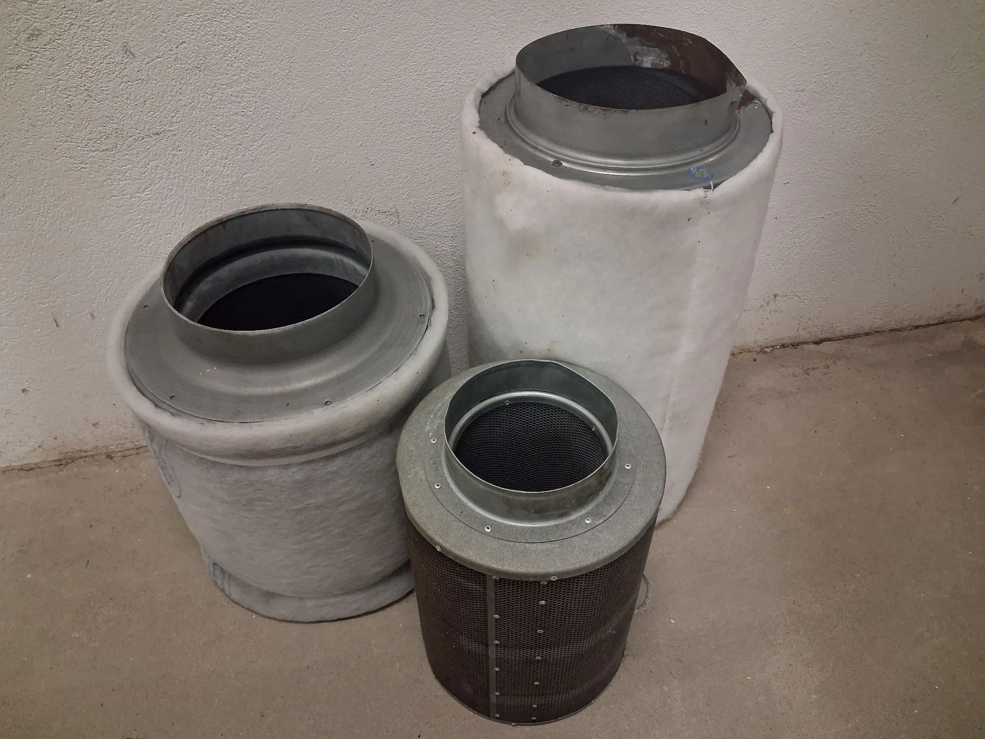 Trzy cylindryczne filtry przemysłowe o różnych rozmiarach i typach wypełnienia, ustawione pionowo na betonowej podłodze na tle białej ściany. Najwyższy filtr po prawej jest owinięty grubą warstwą białego materiału włóknistego. Filtr po lewej jest nieco niższy i ma podobne białe wypełnienie. Najmniejszy filtr z przodu ma ciemną, metalową konstrukcję z drobną siatką.