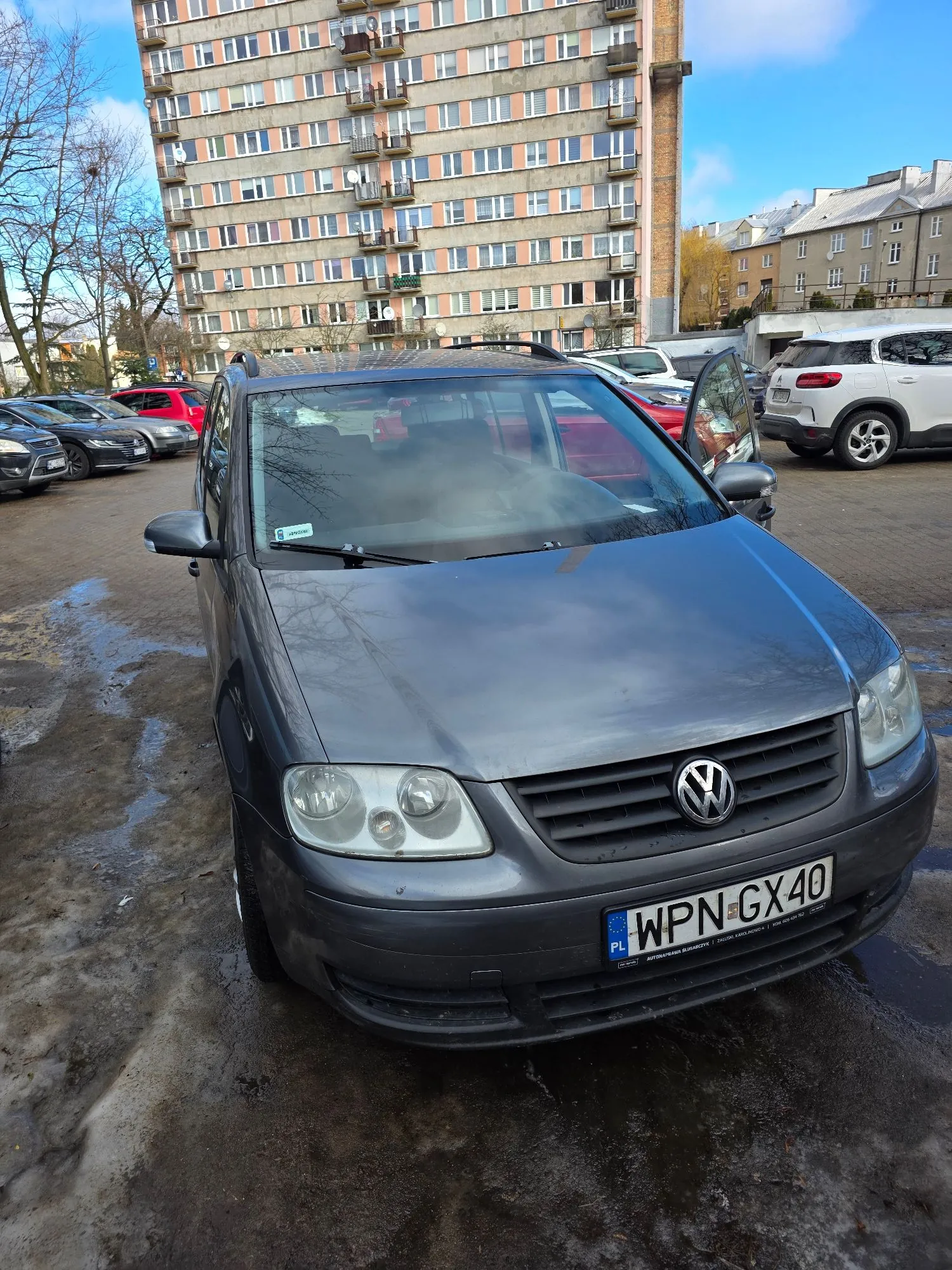 Srebrny samochód marki Volkswagen zaparkowany na błotnistym placu przed wielopiętrowym blokiem mieszkalnym. Na przednim zderzaku widoczna polska tablica rejestracyjna o numerze WPN GX40. W tle znajdują się inne zaparkowane samochody oraz wysoki budynek z licznymi oknami i balkonami.