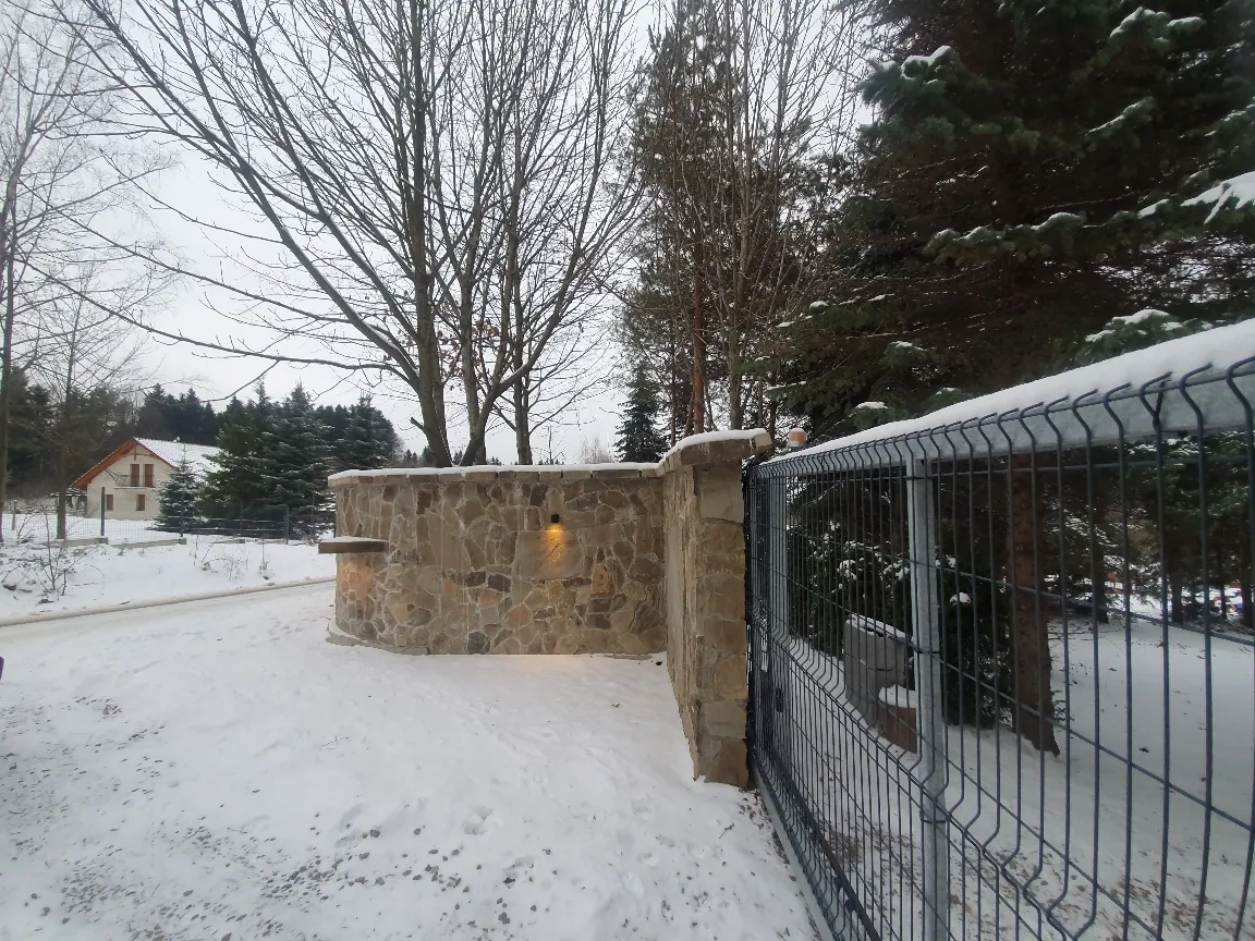 Zimowy krajobraz z kamiennej ściany i metalowego ogrodzenia pokrytych śniegiem. W tle dom z czerwonym dachem, drzewa bez liści i zasneżone iglaste drzewa. Śnieżna droga z odciskami stóp, pochmurne niebo.