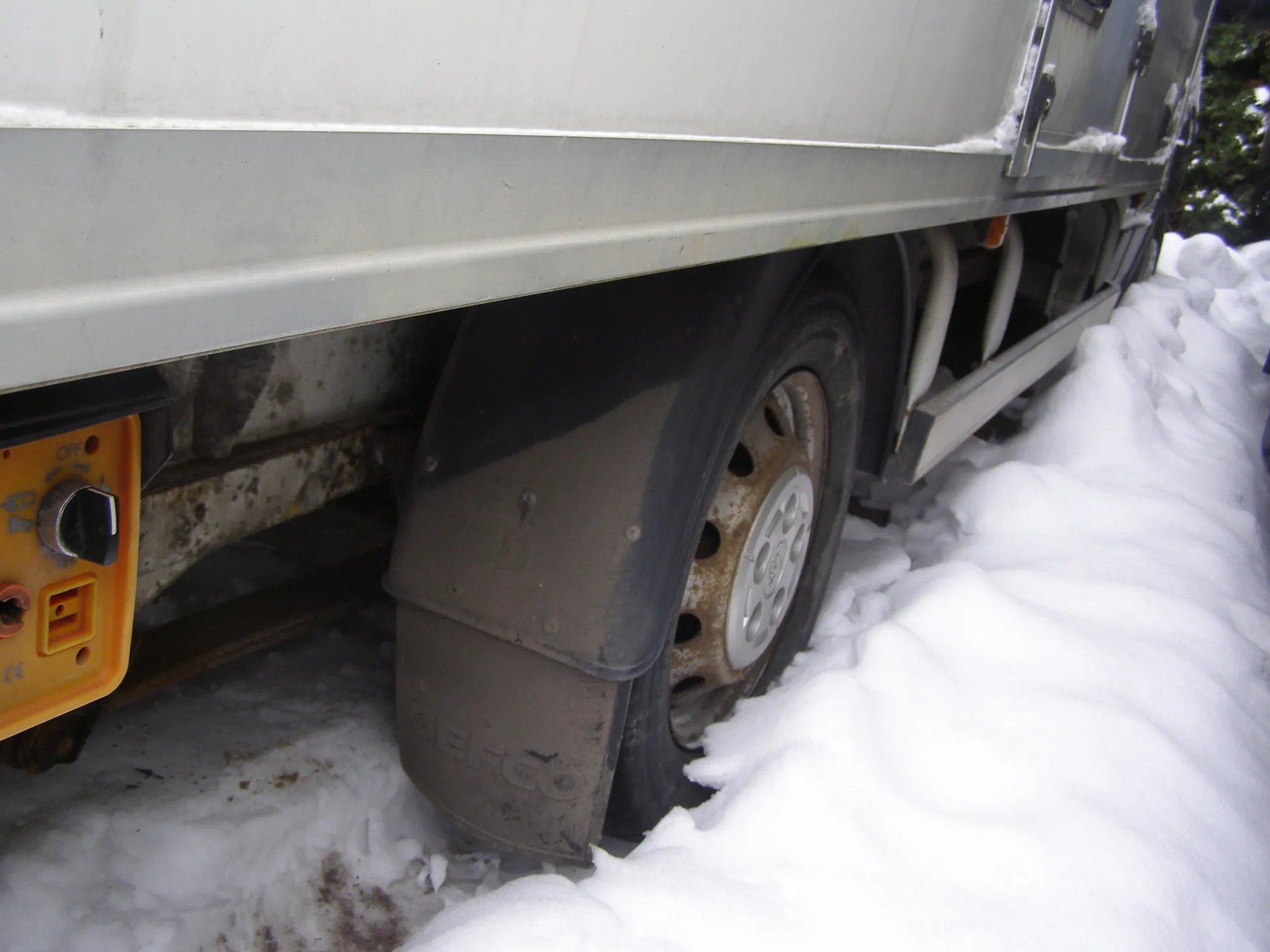 Zbliżenie na tylną część srebrnej ciężarówki z kołem o rdzewnej felgach w śniegu. Brudny błotnik z napisem „GEFCO”, pomarańczowa tablica z gniazdami elektrycznymi. Śnieg pokrywa podłogę i fragmenty pojazdu, widoczne ślady brudu.