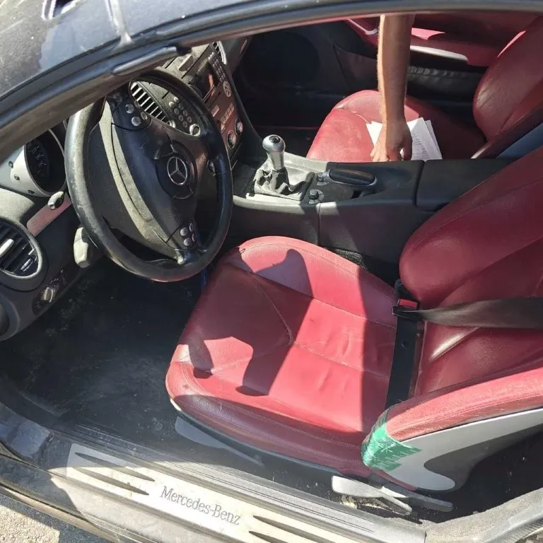 Wnętrze samochodu Mercedes-Benz z czerwonymi skórzany siedzeniami. Widoczna kierownica z logo Mercedes-Benz, deska rozdzielcza, dźwignia zmiany biegów oraz próg drzwi z napisem "Mercedes-Benz". Na siedzeniu pasażera leżą dokumenty, a ręka osoby sięga w ich kierunku. Wnętrze jest dobrze oświetlone, a na podłodze widoczne są ślady zużycia.