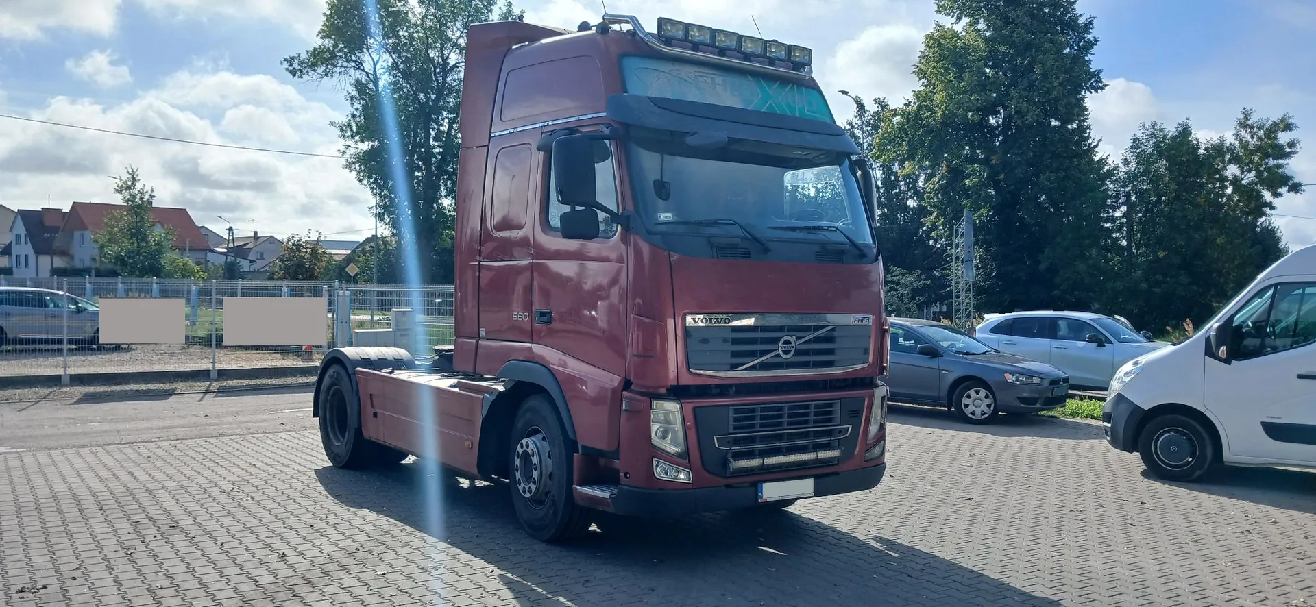 Czerwona ciągnik siodłowy marki Volvo zaparkowany na kostce brukowej. Pojazd posiada dodatkowe oświetlenie na dachu oraz napis 'VOLVO' na przednim grillu. W tle widoczne są zaparkowane samochody osobowe, biały furgon, drzewa oraz zabudowania mieszkalne pod niebem z lekkim zachmurzeniem.
