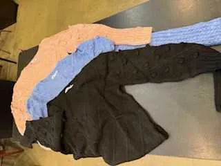 Na szarym stole leżą cztery swetry ułożone w rzędzie. Od lewej: sweter w kolorze brzoskwiniowym, niebieski sweter z dzianiny, jasnoniebieski sweter z dzianiny oraz czarny sweter z guzikami. Tło składa się z częściowo widocznego drewnianego podłogowego i szarego powierzchni stołu.