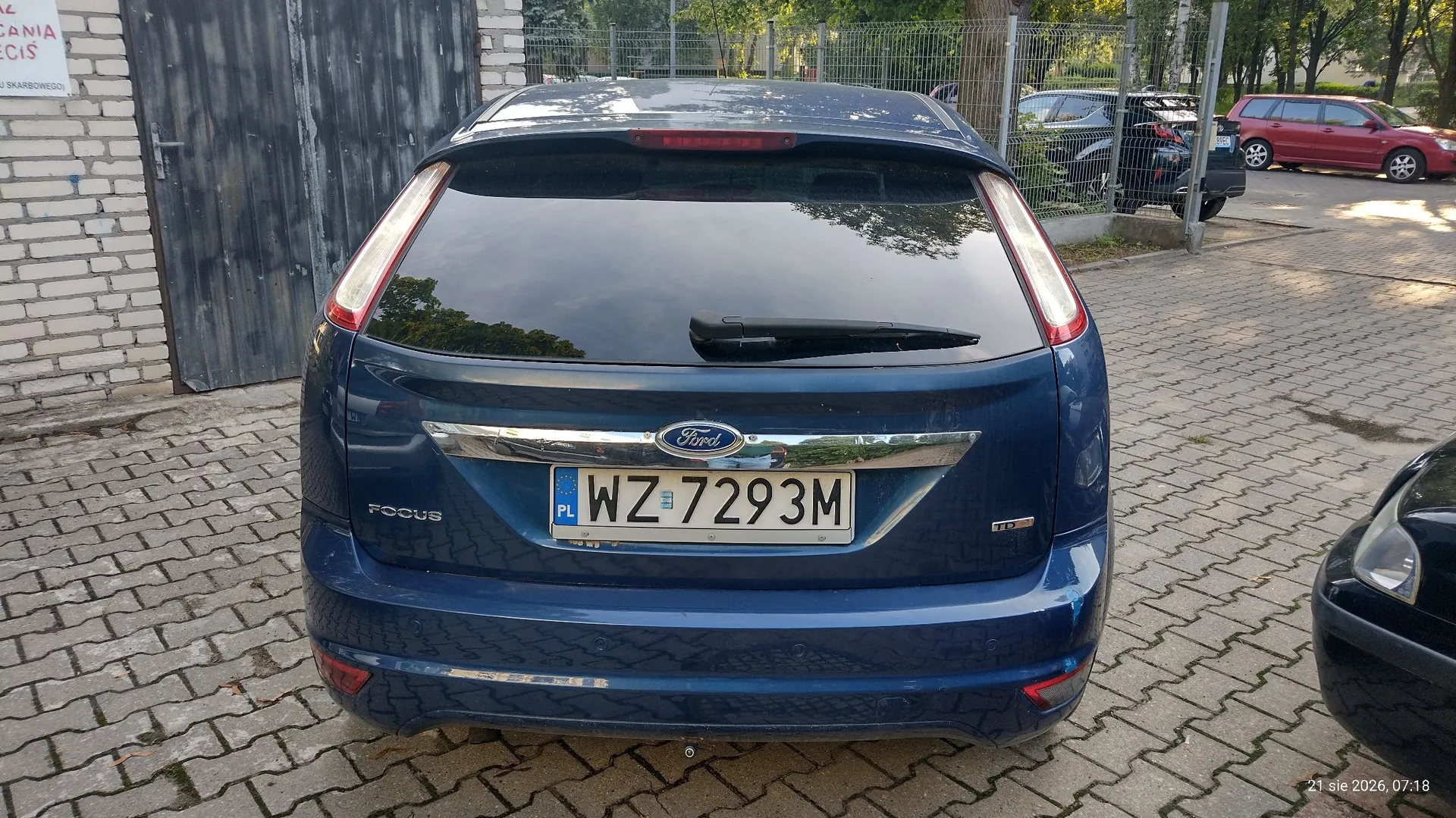 Tylny widok niebieskiego samochodu marki Ford Focus zaparkowanego na kostce brukowej. Na klapie bagażnika widoczna jest tablica rejestracyjna o numerze WZ 7293M oraz logo Ford. W tle widać biały mur, szarą bramę oraz inne zaparkowane samochody za ogrodzeniem z siatki.