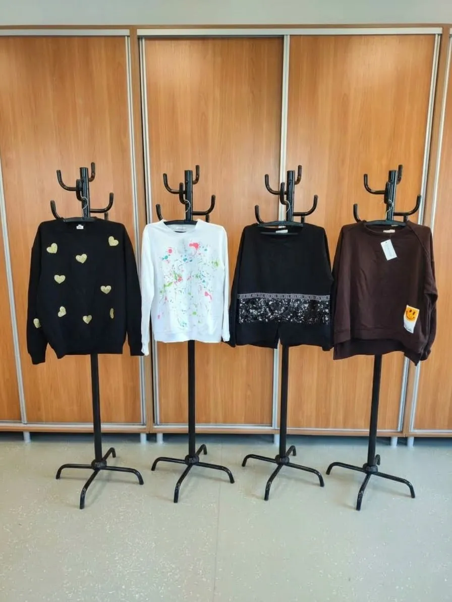 Cztery czarne i białe bluzy wyeksponowane na czarnych stojakach przed drewnianymi szafami. Od lewej: czarna bluza z małymi złotymi sercami, biała bluza z kolorowym nadrukiem kwiatowym, czarna bluza z ozdobnym pasem koronki na dole oraz brązowa bluza z małym żółtym elementem na rękawie.