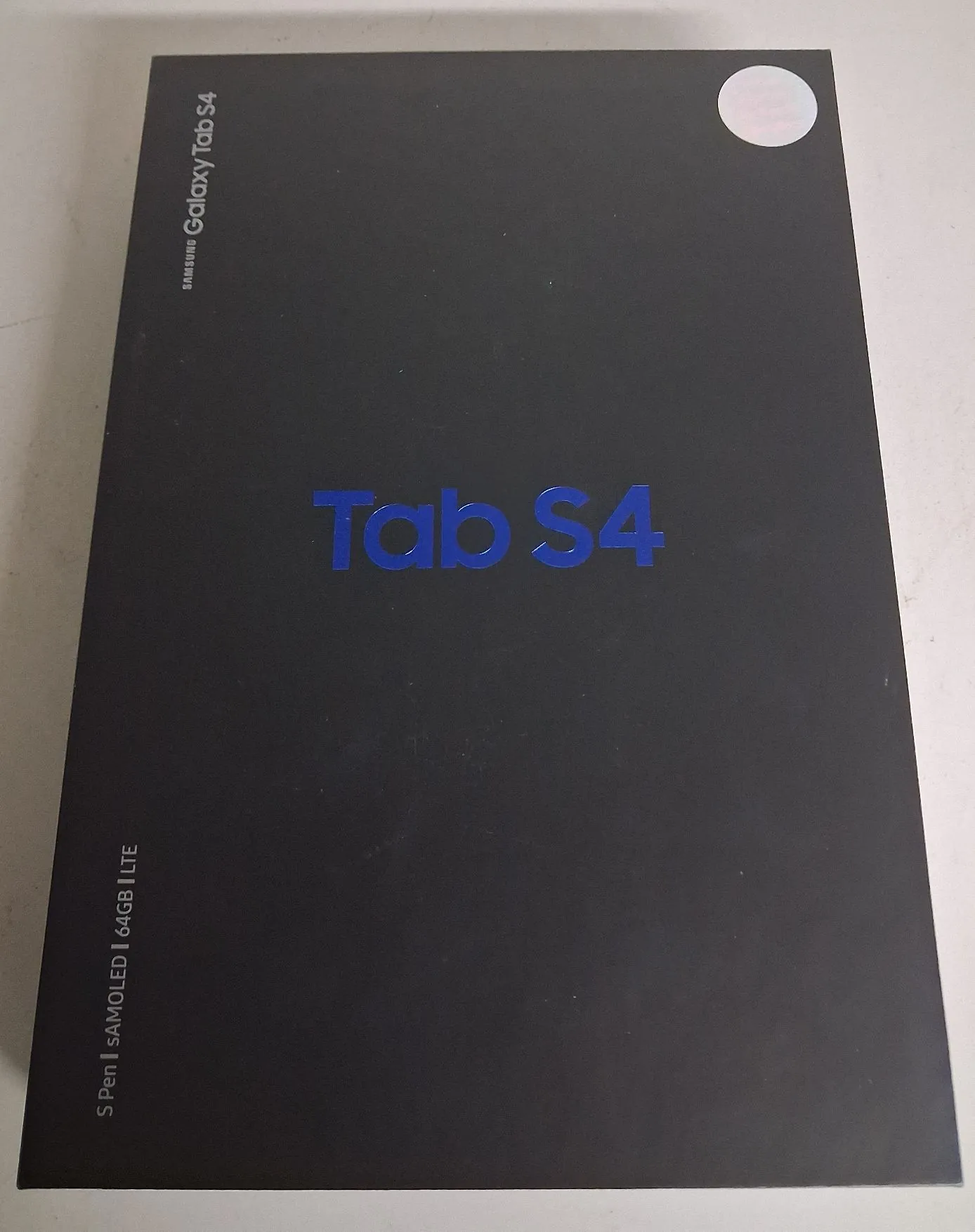 Czarna karta opakowania tabletu Samsung Galaxy Tab S4. Na środku znajduje się duży, niebieski napis 'Tab S4'. W górnej lewej części widnieje pionowy napis 'SAMSUNG Galaxy Tab S4', a w dolnej lewej części pionowy napis 'S Pen | SAMOLED | 64GB | LTE'. W prawym górnym rogu znajduje się okrągła, biała naklejka.