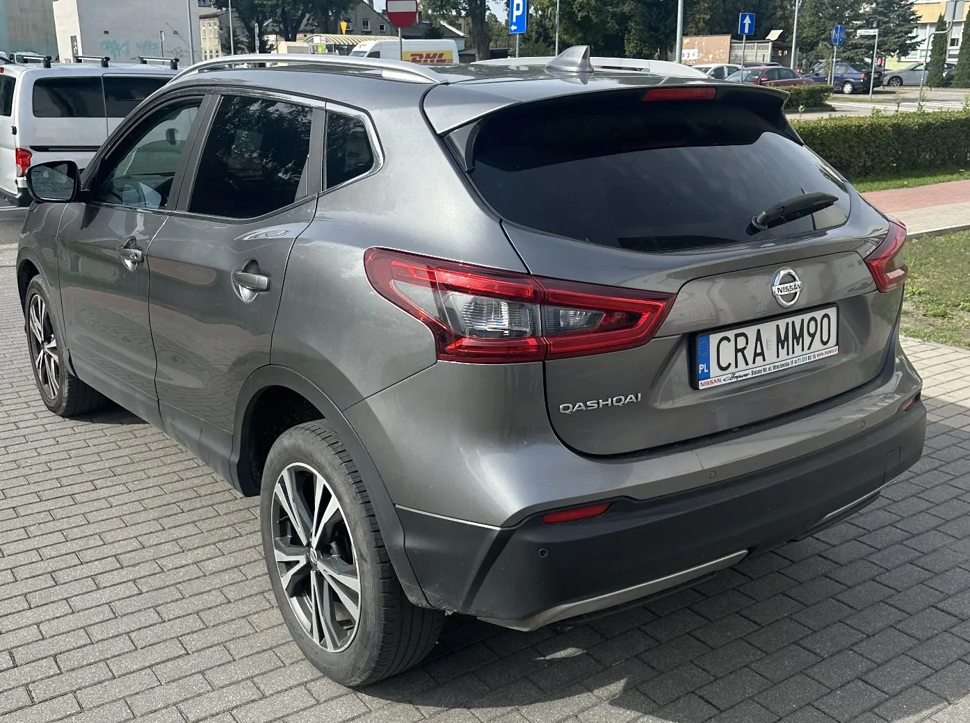 Srebrnoszary samochód marki Nissan Qashqai zaparkowany na kostce brukowej. Widok z tyłu i z boku auta. Na klapie bagażnika znajduje się logo Nissana, napis Qashqai oraz polska tablica rejestracyjna o numerze CRA MM90. W tle widoczne są inne zaparkowane samochody, znaki drogowe oraz drzewa.