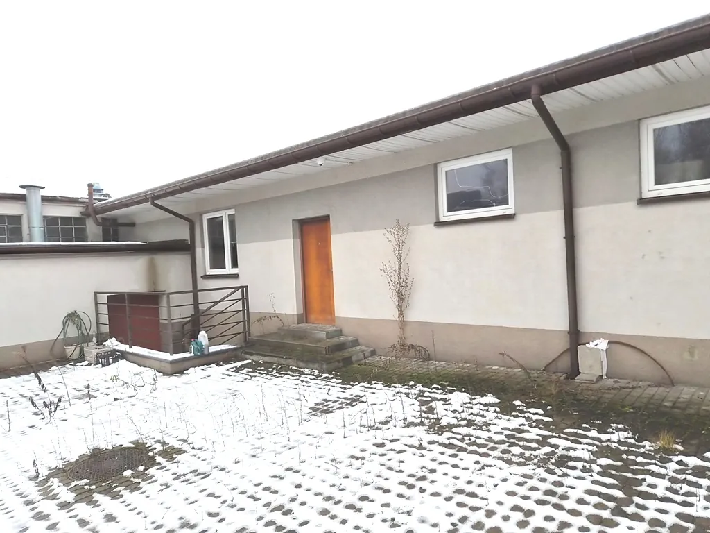Zdjęcie przedstawia zewnętrzną ścianę parterowego budynku o jasnej elewacji z brązowym rynkiem i dachem. W ścianie znajdują się dwa białe okna oraz jedne drewniane, pomarańczowe drzwi wejściowe z betonowymi schodami. Po lewej stronie widoczny jest niski murek z metalową barierką i czerwonym elementem. Przed budynkiem znajduje się teren pokryty cienką warstwą śniegu, spod której prześwituje ziemia i roślinność.