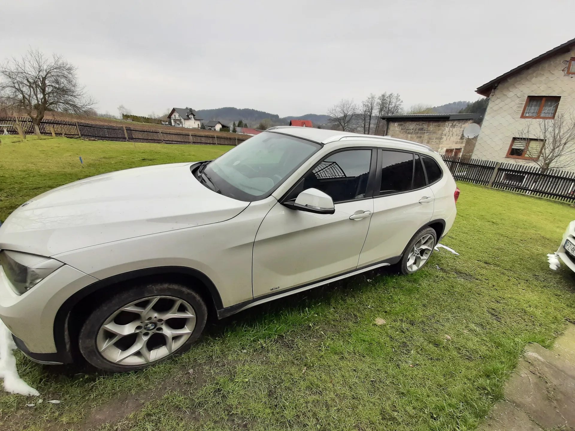 Biały SUV marki BMW zaparkowany na trawie. W tle widoczne domy, ogrodzenie, drzewa bez liści oraz wzgórza. Niebo jest zachmurzone. Po prawej stronie częściowo widoczna tablica rejestracyjna z literami 'PZ'. Na trawie znajdują się resztki śniegu.
