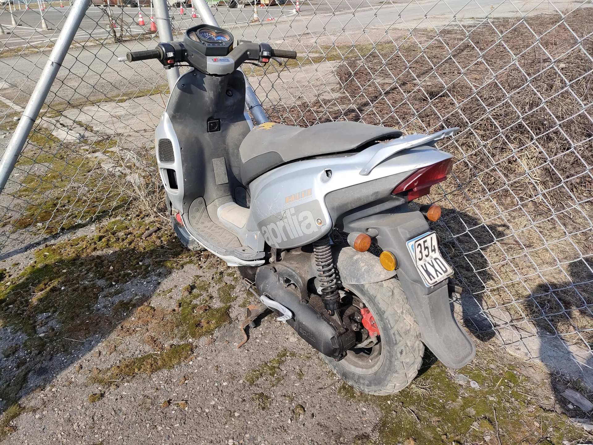 Srebrno-czarny skuter marki Aprilia zaparkowany na żwirowym podłożu z plamami mchu, obok metalowego ogrodzenia siatkowego. Widoczna tablica rejestracyjna z numerem 354 KXD, tylna część skutera z czerwonym światłem i pomarańczowymi kierunkowskazami. W tle fragment drogi z pomarańczowymi stożkami.