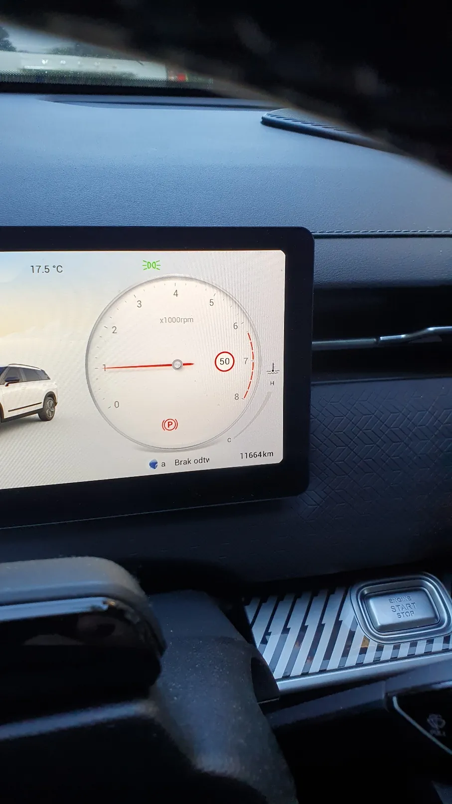Zdjęcie wnętrza samochodu przedstawiające cyfrowy wyświetlacz deski rozdzielczej. Na ekranie widoczny jest obrotomierz z wartością x1000rpm, temperaturą 17.5 °C, aktualnym przebiegiem 11664 km oraz ikoną biegu P (parkowanie) i napisem 'Brak odtw'. W prawym dolnym rogu widoczny jest srebrny przycisk 'ENGINE START STOP'.