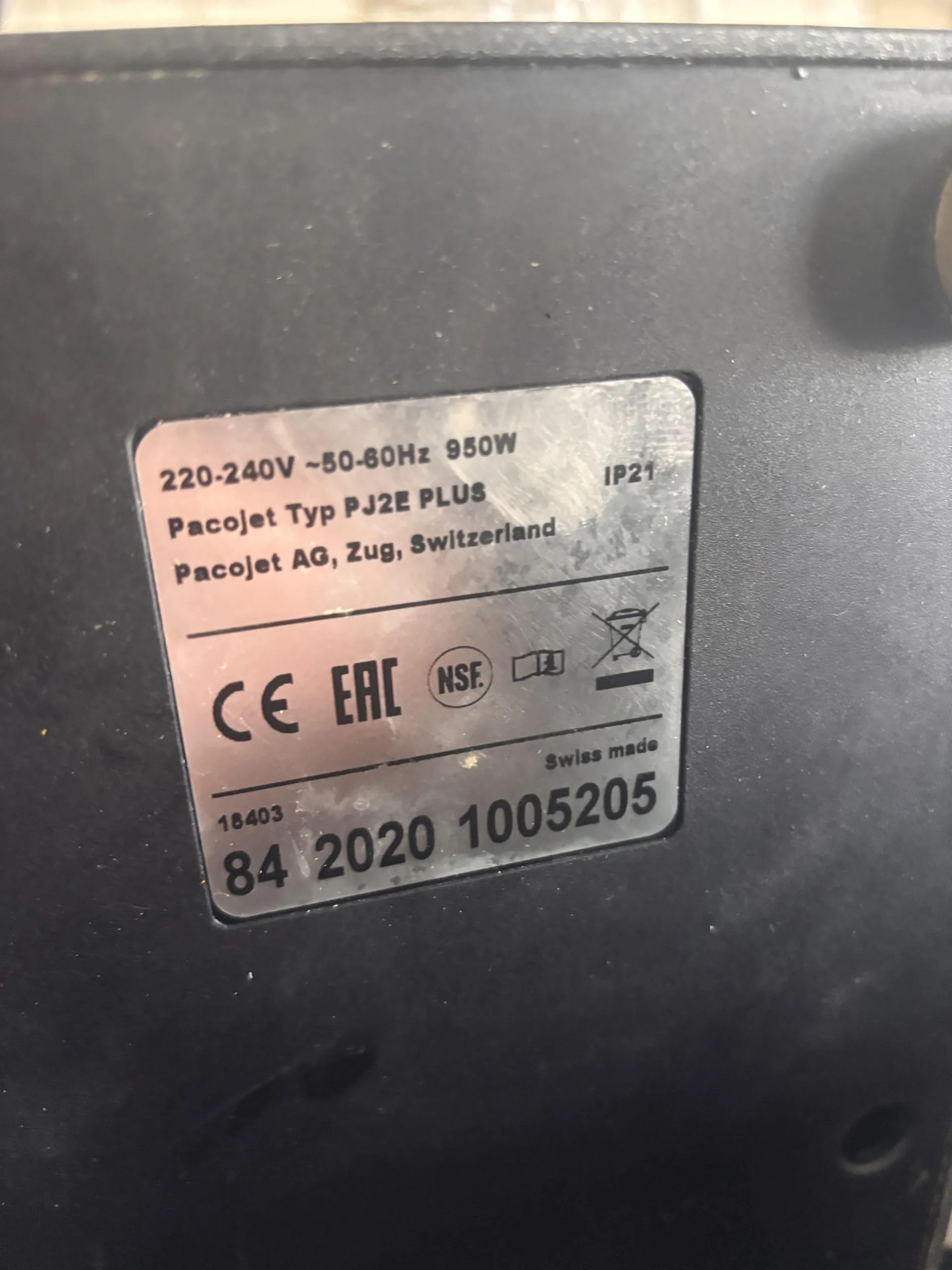 Czarna powierzchnia z metalową tabliczką znamionową. Na tabliczce widoczny tekst: 220-240V ~50-60Hz 950W, Pacojet Typ PJ2E PLUS, Pacojet AG, Zug, Switzerland, IP21. Poniżej znaki certyfikacyjne: CE, EAC, NSF, ikona książki, symbol zakazanego wyrzucania do śmieci. Dalsze informacje: Swiss made, 18403, 84 2020 1005205. Tekst na tabliczce jest ułożony poziomo, czytelny.