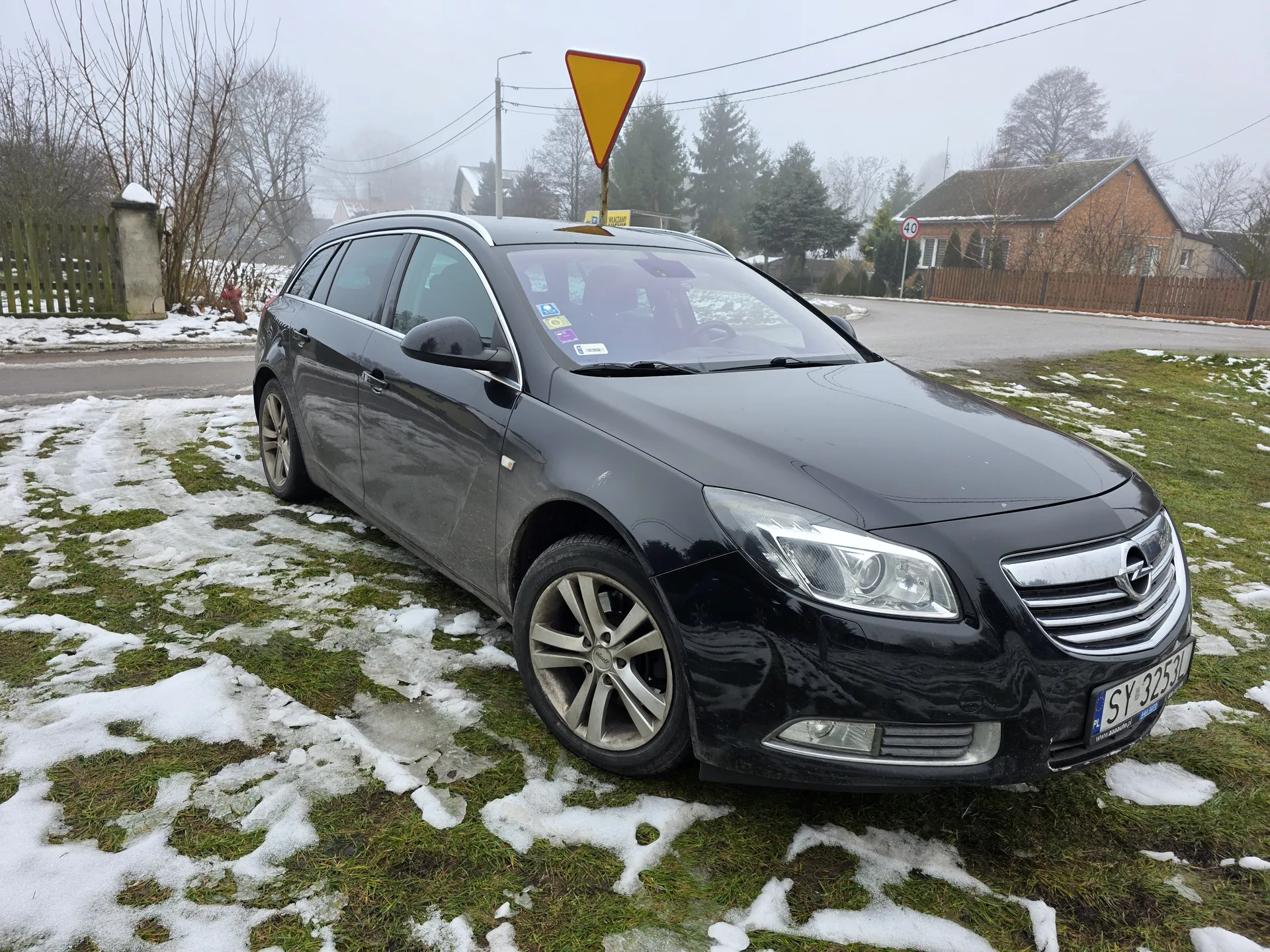 Czarny samochód osobowy typu kombi marki Opel zaparkowany na trawie z resztkami śniegu. Na przednim szybie widoczne są naklejki, a na tablicy rejestracyjnej napis "SY 3253L" z oznaczeniem "PL". W tle żółty trójkątny znak drogowy ostrzegawczy, domy, drzewa bez liści, ogrodzenie oraz droga z tablicą prędkości "40". Pogoda mglista, na ziemi śnieg i zamarznięta trawa.