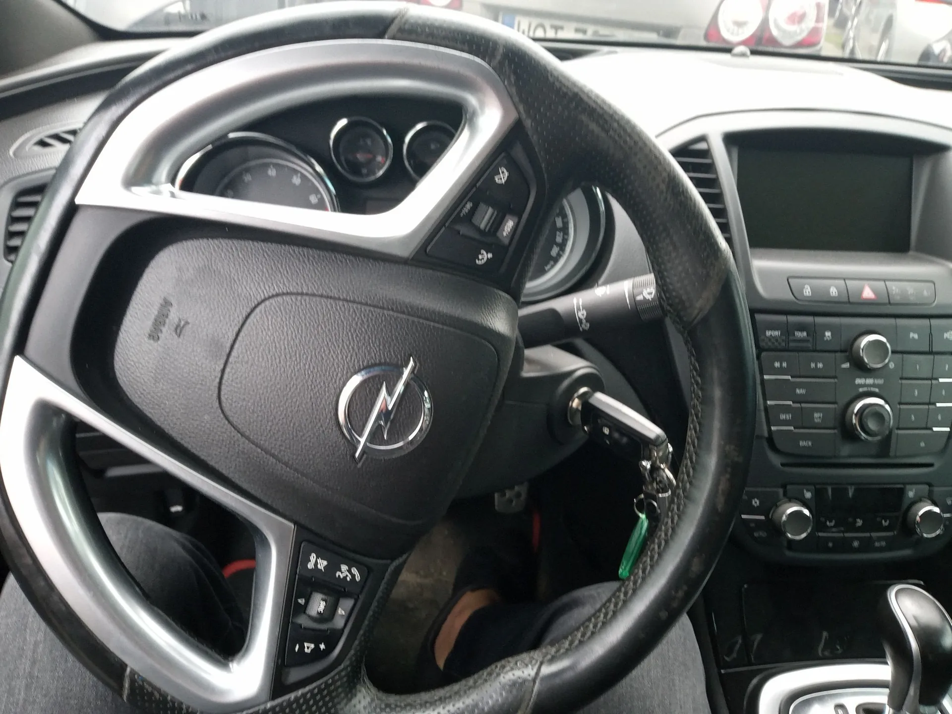 Widok z wnętrza samochodu marki Opel, skupiony na kierownicy i desce rozdzielczej. Kierownica jest czarna, obszyta skórą z srebrnymi wstawkami i logo Opela w centrum. Po prawej stronie widoczny jest panel centralny z ekranem, przyciskami sterowania radiem i klimatyzacją oraz lewarem zmiany biegów. W stacyjce znajduje się włożony kluczyk z zielonym brelokiem.