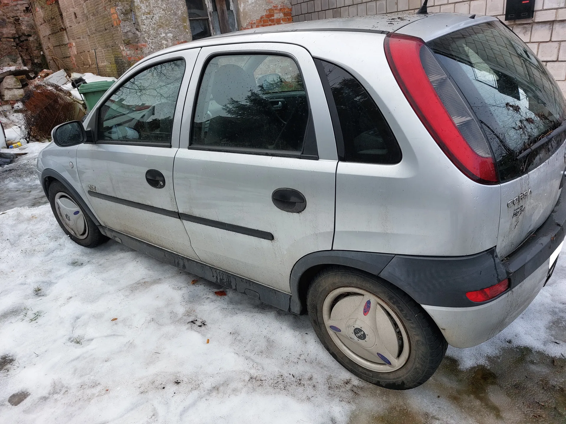 Zdjęcie przedstawia srebrny samochód Opel Corsa zaparkowany na śnieżnej powierzchni. Pojazd ma czarne elementy na zderzakach i nadkółach, brudne boki oraz felgi z niebiesko-czerwonymi naklejkami. W tle widoczne są zniszczone budynki z cegły i betonu, zielony pojemnik na śmieci oraz śnieg na ziemi. Na tylnym zderzaku widnieje napis 'CORSA'.