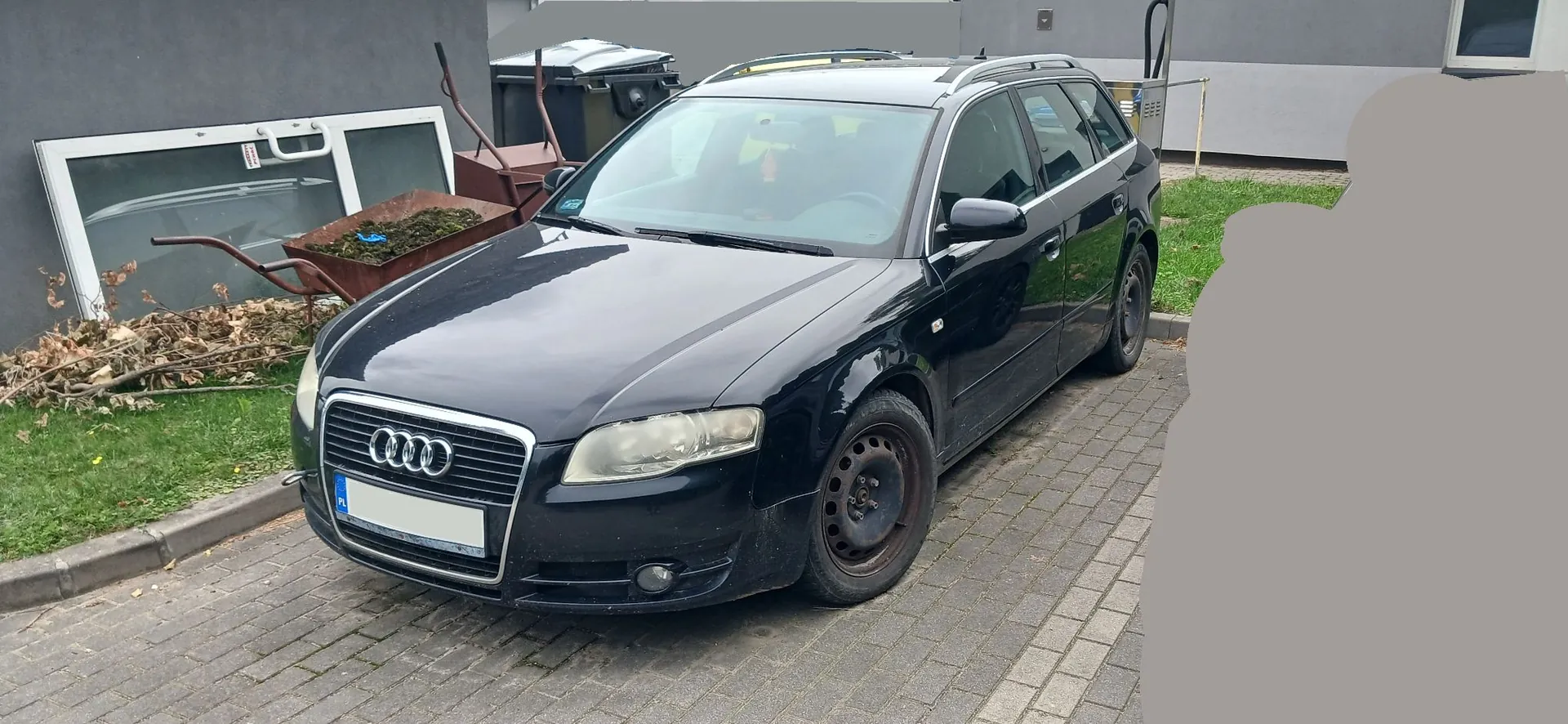Czarny samochód marki Audi w nadwoziu kombi zaparkowany na kostce brukowej obok trawnika. W tle widoczny jest szary budynek, czarny kontener na śmieci oraz metalowa taczka wypełniona liśćmi i gałęziami. Po lewej stronie leży sterta suchych gałęzi i fragment ramy okiennej.