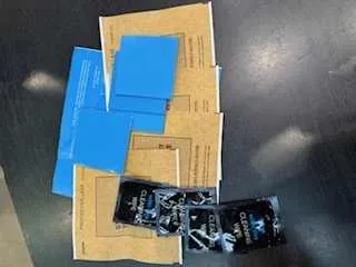 Na ciemnym, matowym stole ułożone są brązowe kartonowe opakowania, niebieskie arkusze papieru oraz dwa czarne opakowania z nieczytelnym tekstem i grafikami.