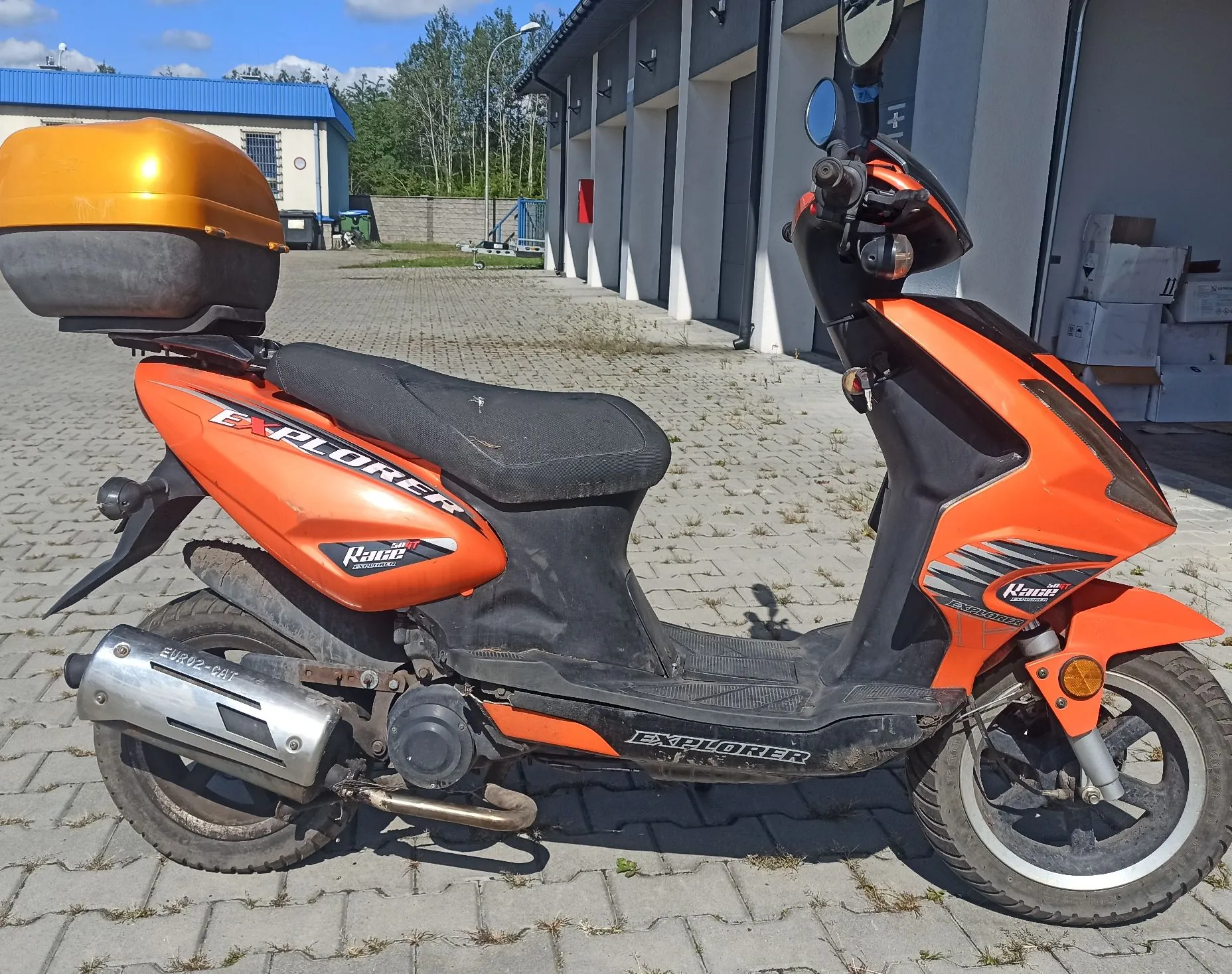 Pomarańczowy skuter marki Explorer zaparkowany na kostce brukowej przed białym budynkiem. Skuter posiada czarne siedzenie, pomarańczowy kufer zamykany na klucz zamontowany z tyłu oraz srebrny tłumik wydechu. Na boku skutera widnieją napisy 'EXPLORER' oraz logo 'Kanza'.
