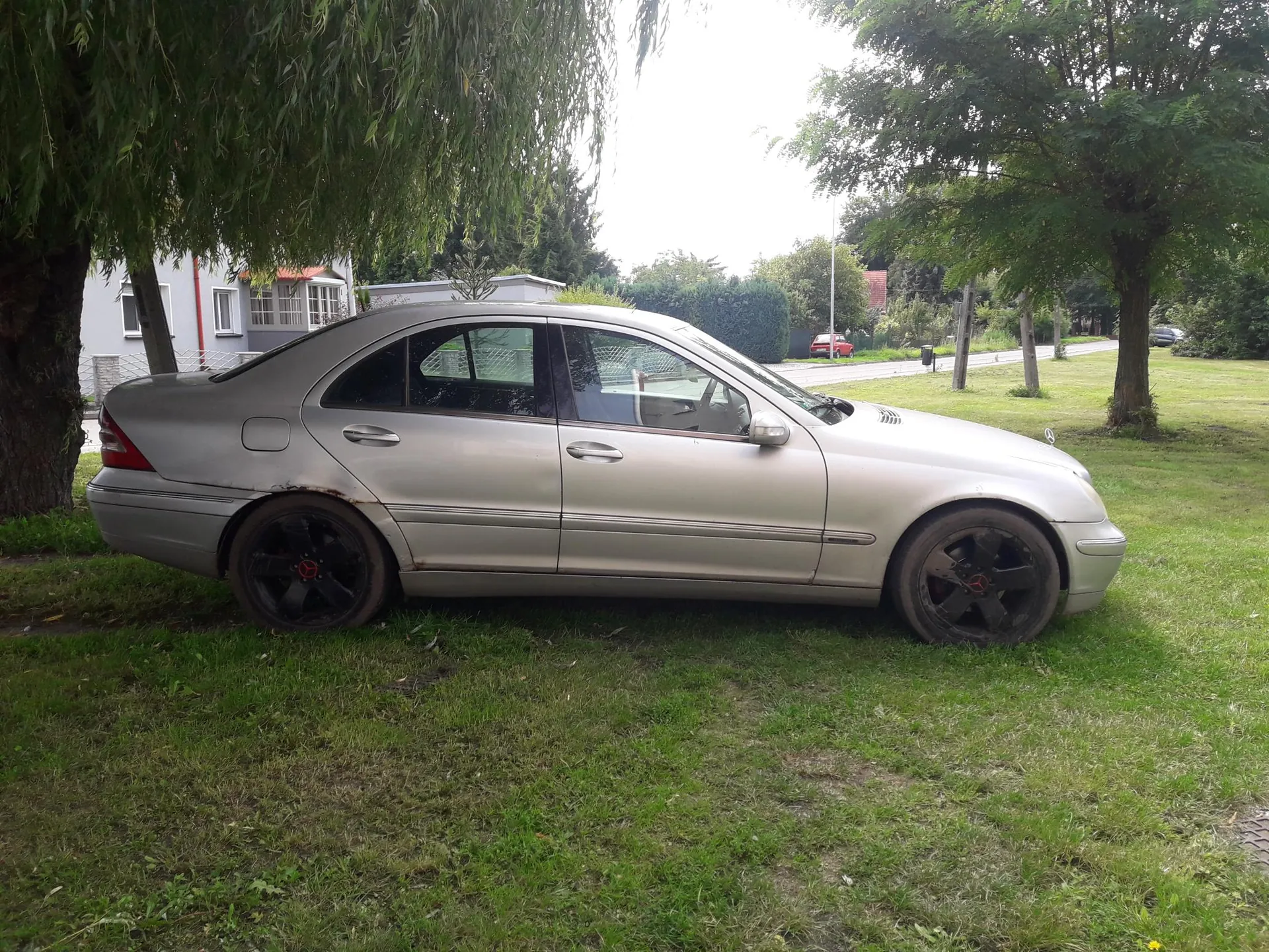 Srebrny samochód osobowy typu sedan marki Mercedes zaparkowany na trawie. Pojazd ma czarne felgi z czerwonym logo w środku, widoczne są ślady brudu na karoserii i kołach. W tle drzewa, biały dom z czerwonymi elementami, ogrodzenie, droga oraz czerwony samochód w oddali. Trawa jest zielona z miejscami ubytków.