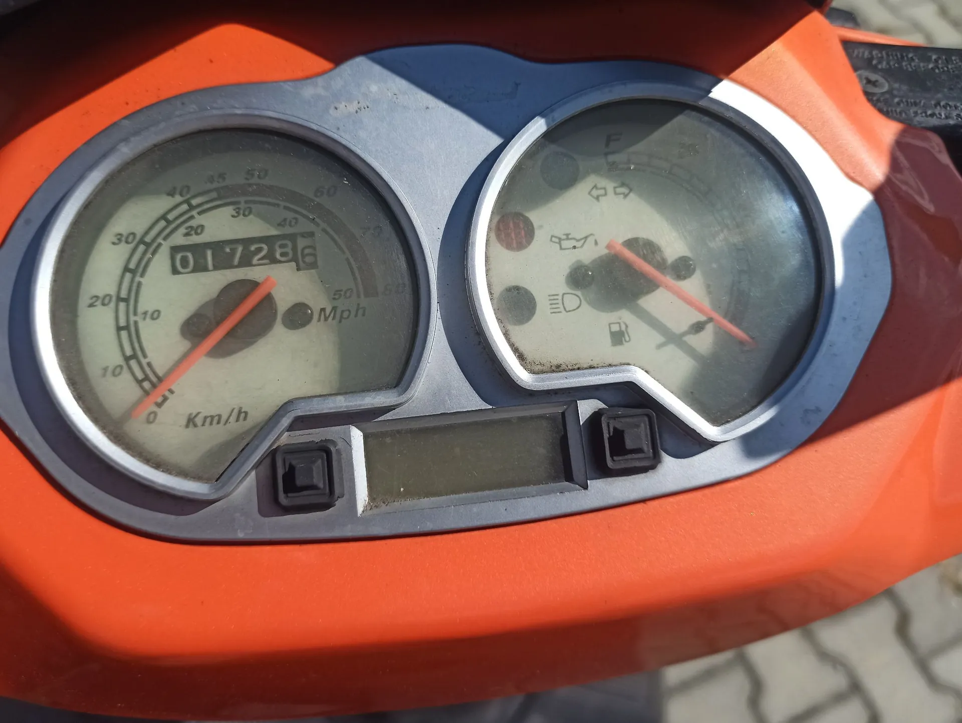 Zbliżenie na deskę rozdzielczą pomarańczowego motocykla. Widoczne są dwa okrągłe wskaźniki w srebrnej obudowie. Lewy wskaźnik to prędkościomierz z podziałką w km/h i Mph, z licznikiem kilometrów wyświetlającym wartość 01728. Prawy wskaźnik zawiera wskaźnik poziomu paliwa oraz kontrolki ostrzegawcze, w tym ikonę silnika i świateł. Poniżej zegarów znajduje się mały, prostokątny wyświetlacz LCD oraz dwa czarne przyciski.