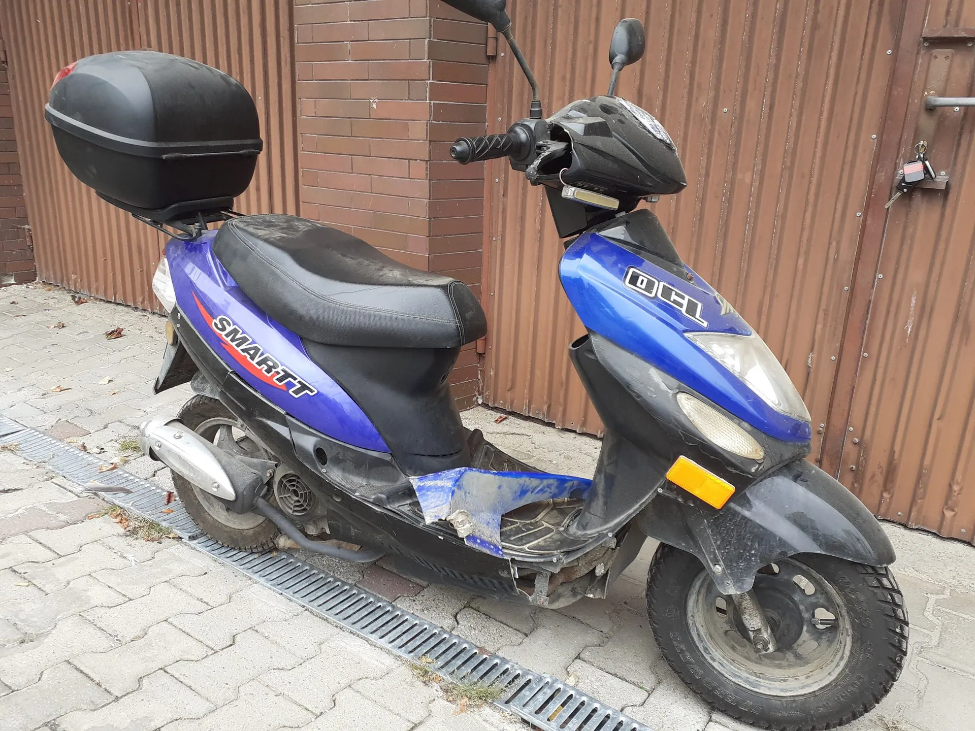 Niebieski skuter marki Smartt z czarnym siedzeniem i czarnym kufrem z tyłu. Pojazd stoi na kostce brukowej przed brązową, metalową bramą i ceglaną ścianą. Na boku skutera widnieje biały napis „SMARTT”, a na przedniej owiewce napis „DCL”. Skuter posiada widoczne uszkodzenia plastikowych elementów w dolnej części nadwozia.