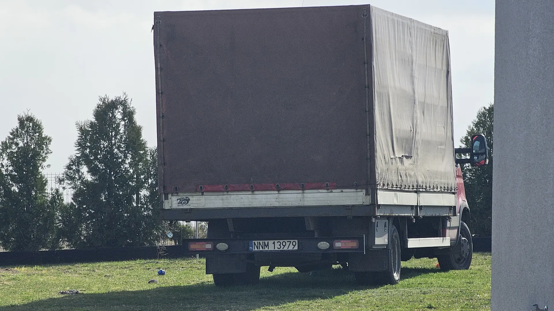 Z tyłu widoczna ciężarówka z przyczepą o brązowym kolorze i szarym tarpem po prawej stronie. Tablica rejestracyjna NNM 13979 umieszczona na tylnej części pojazdu. W tle drzewa, trawa, fragment ogrodzenia oraz budynek po prawej stronie. Niebo jest jasne.