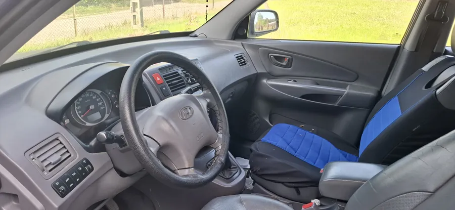 Wnętrze samochodu z widoczną kierownicą marki Hyundai, zegarami samochodowymi, konsolą środkową z przyciskami i wentylacją. Siedzenia pasażerskie są niebiesko-czarne, na siedzeniu pasażera leży czarna torba. Za przednią szybą widoczny jest trawnik i ogrodzenie. Wnętrze posiada szare i czarne elementy wykończenia.