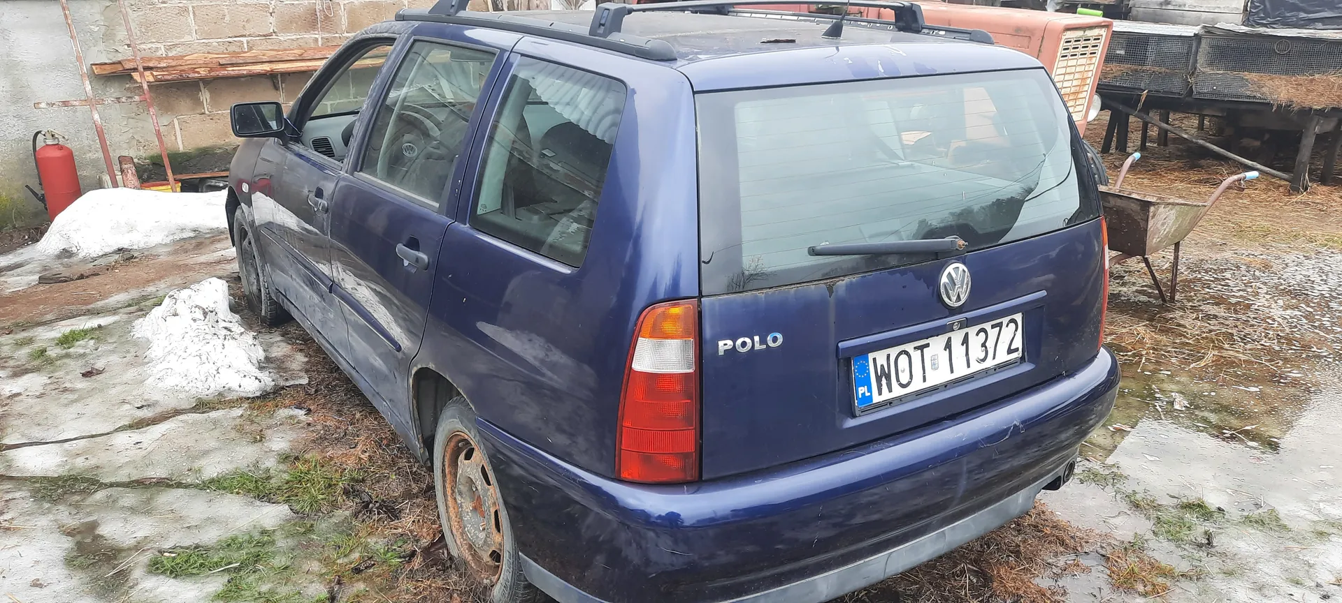 Niebieski Volkswagen Polo z tablicą rejestracyjną WOT 11372 (PL), widok z tyłu. Samochód z bagażnikiem na dachu stoi na betonowej powierzchni z resztkami śniegu i trawą, zardzewiałe koło tylne. W tle ceglana ściana, wózek ogrodniczy, gaśnica i konstrukcje z siatki. Napisy "POLO" i logo VW.